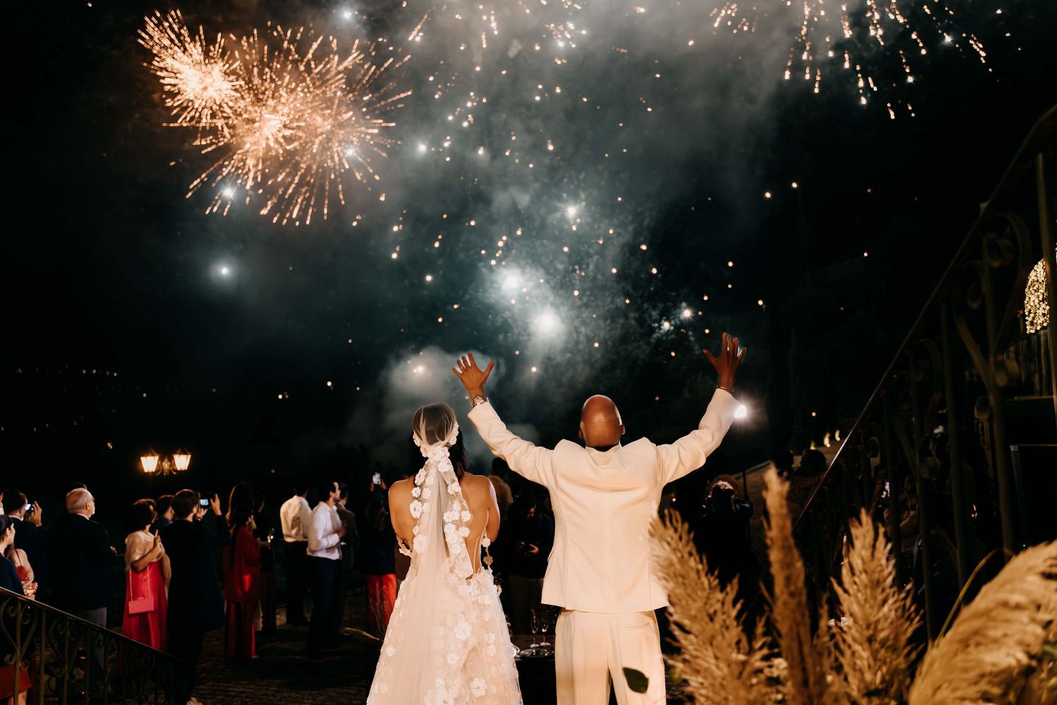 A group watches a dazzling fireworks display bursting in a dark night sky while standing near illuminated trees.