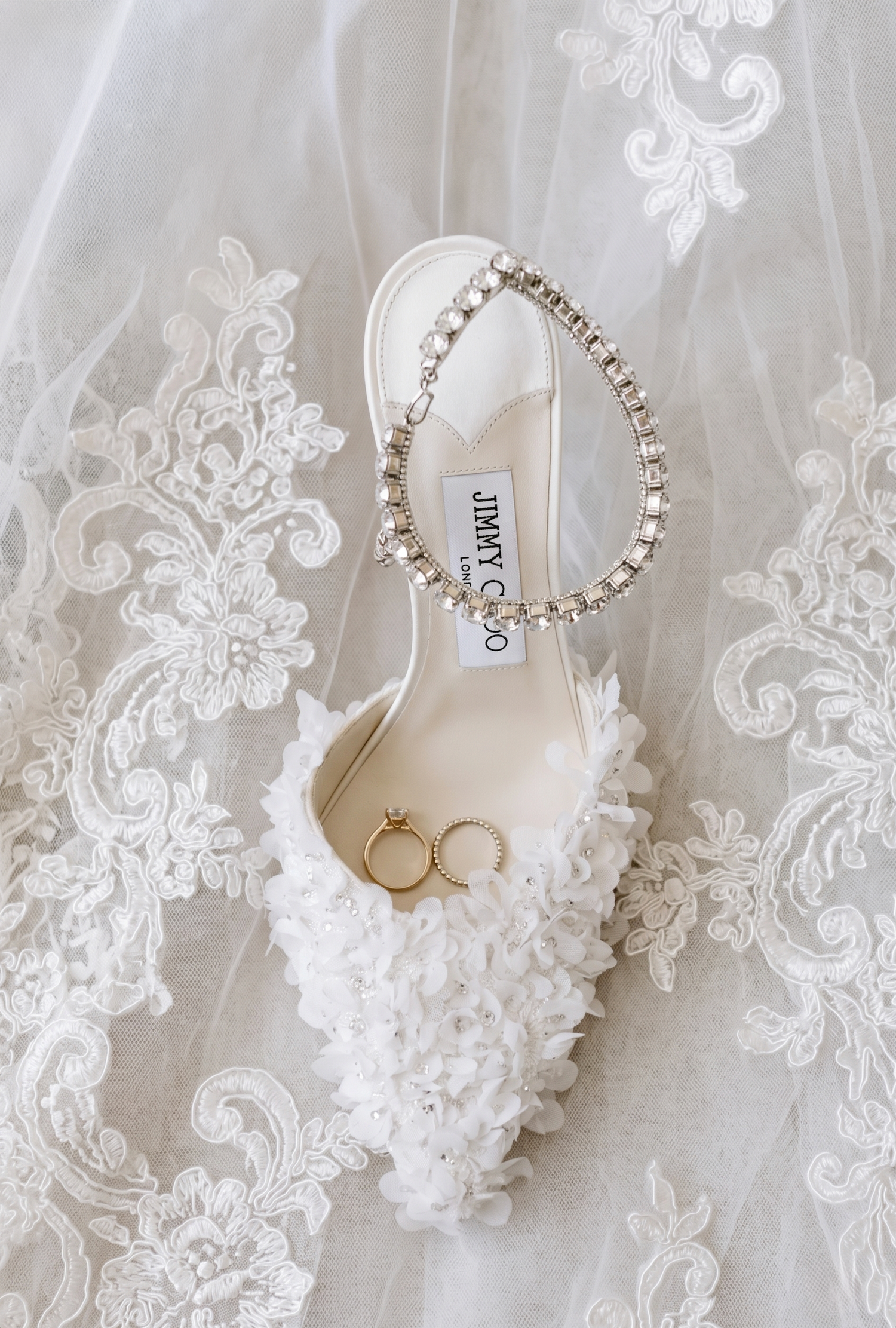 Jimmy Choo white floral bridal heel with crystal ankle strap and gold wedding rings resting on lace wedding veil.
