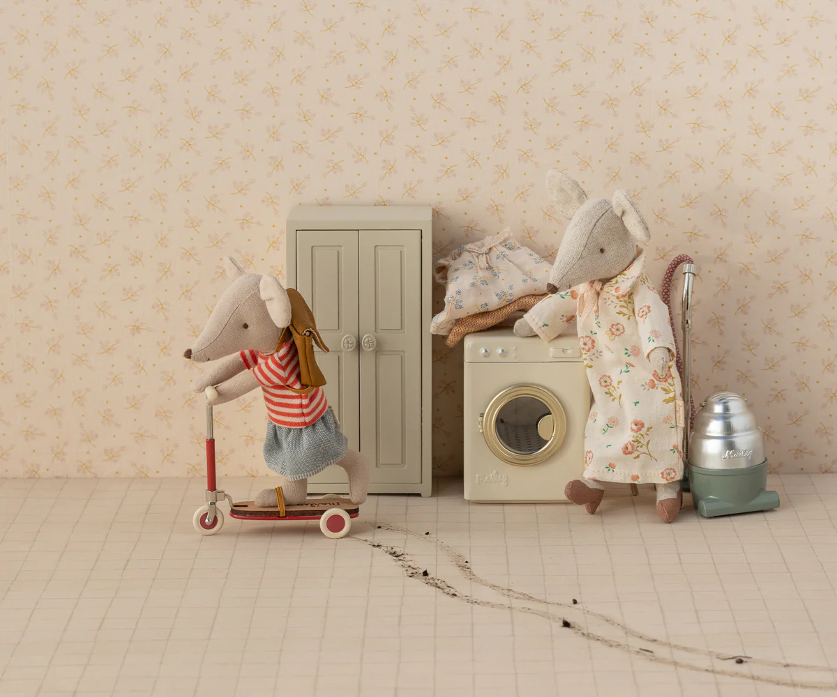 Toy mice doing laundry in miniature scene with washing machine, door, and scooter against beige wallpaper.