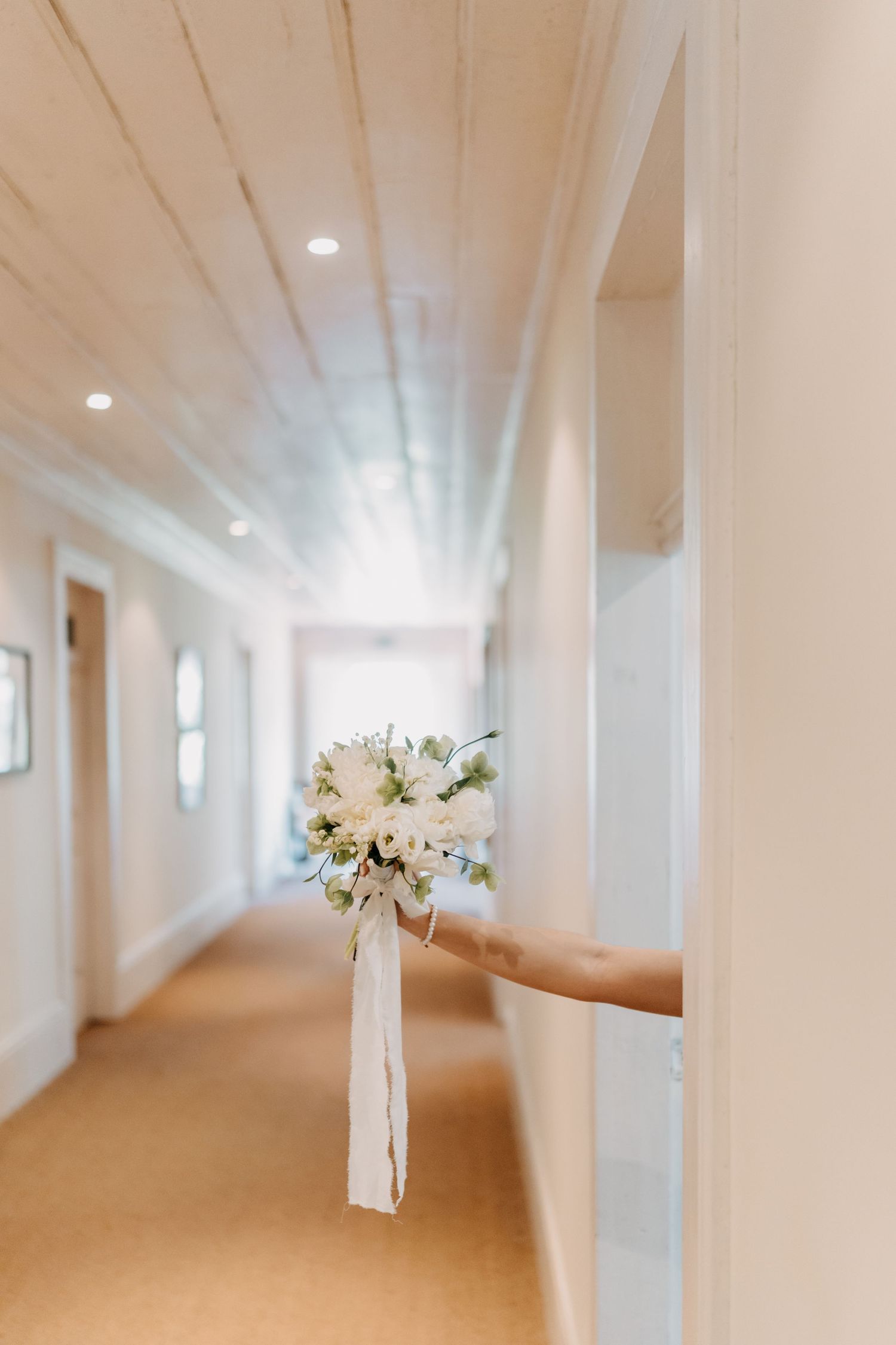 Long bright corridor with white pillars, carpeted beige floor and recessed lighting leads to floral arrangements at the end.