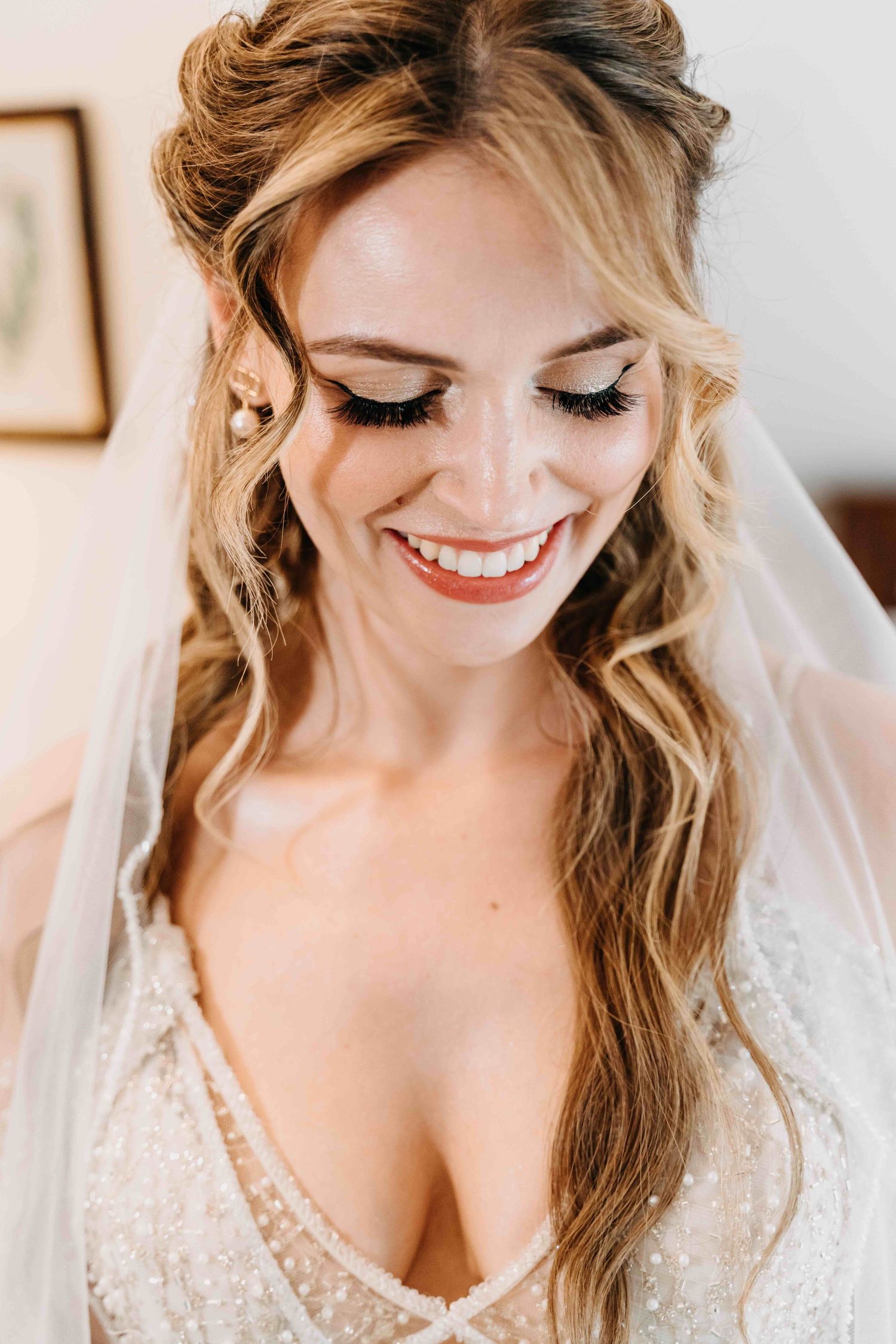 A bride in a lace wedding dress and veil smiles while looking down at her bouquet with soft curled hair.