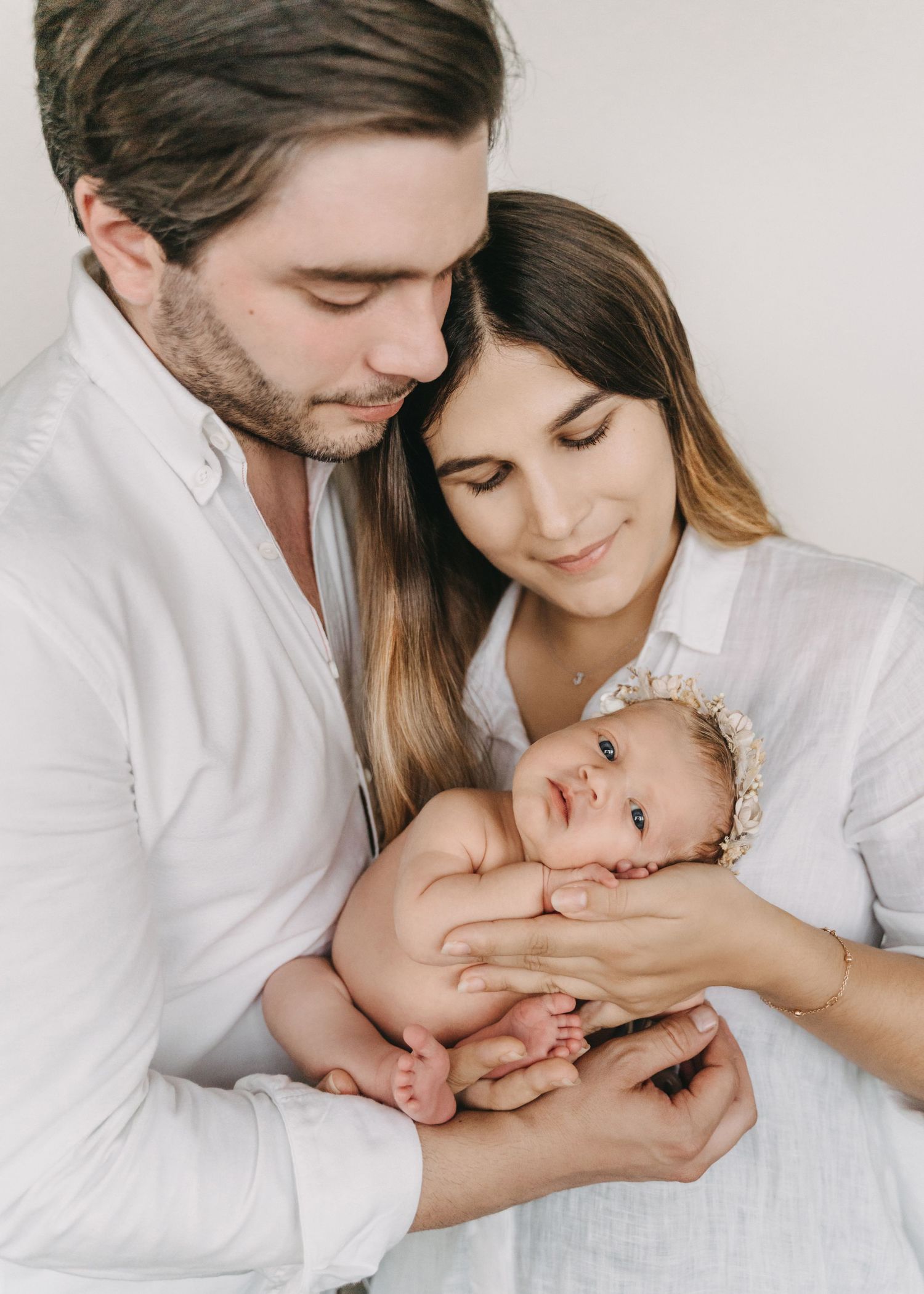Intimate family moment with newborn baby wearing lace bonnet while being held in white room.