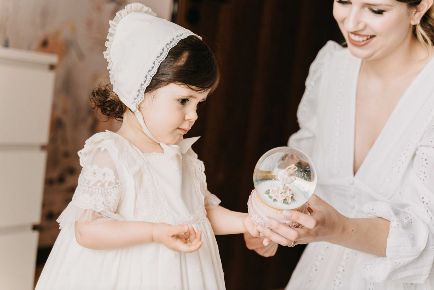 A small child in a white dress and bonnet looks at a glass ornament during what appears to be a wedding preparation.