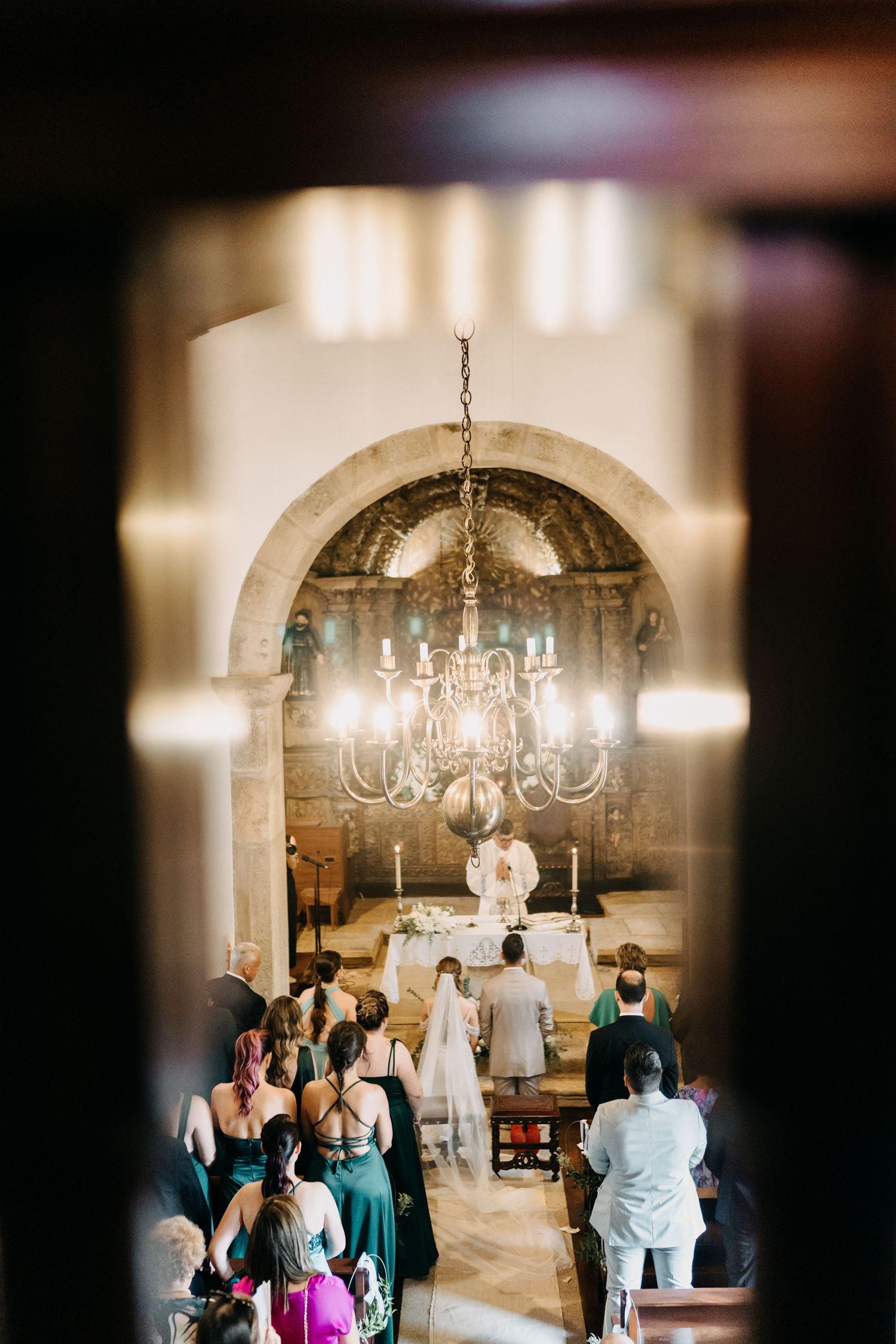 Romantic wedding ceremony in a stone chapel with a chandelier casting warm light over the gathered guests.