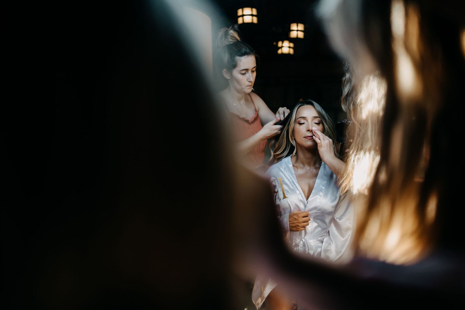 Through a blurred foreground, a bride gets her hair and makeup done in preparation for her wedding day.