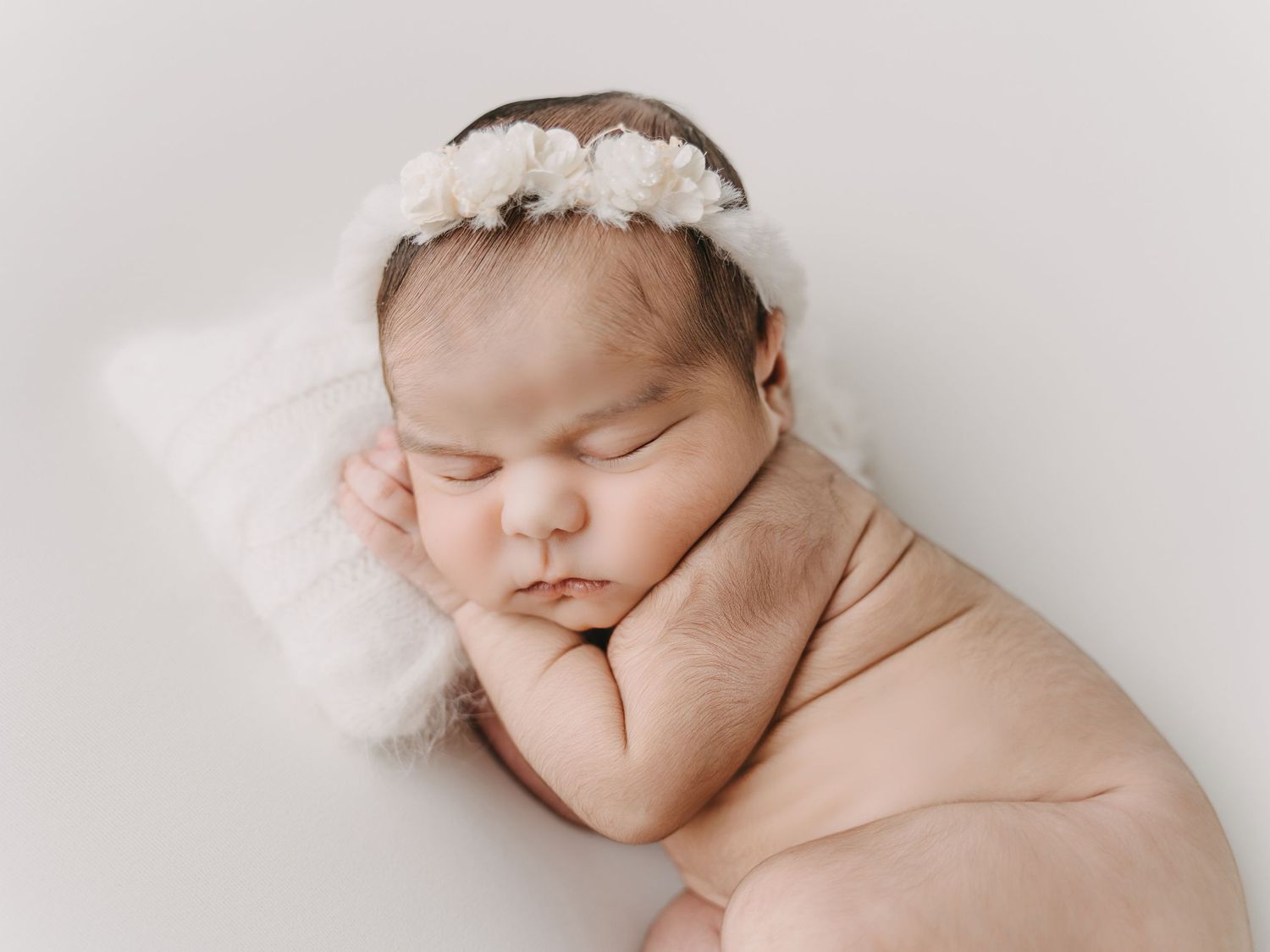 Peaceful sleeping newborn wearing delicate white lace headband resting on soft white pillow in studio portrait.