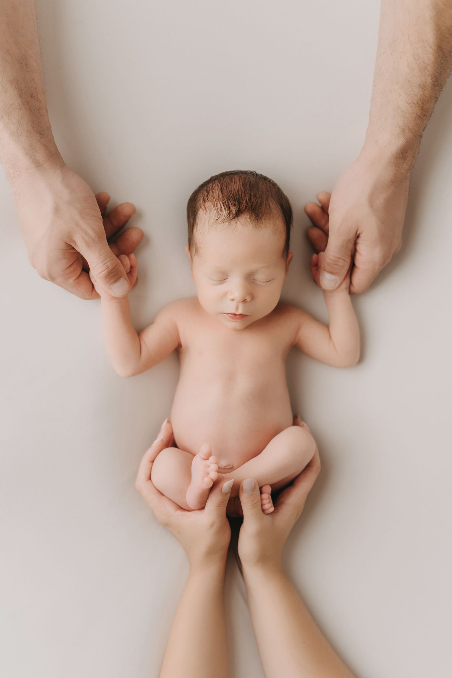 Gentle hands hold a sleeping newborn infant lying on a soft white background in a minimalist studio portrait.