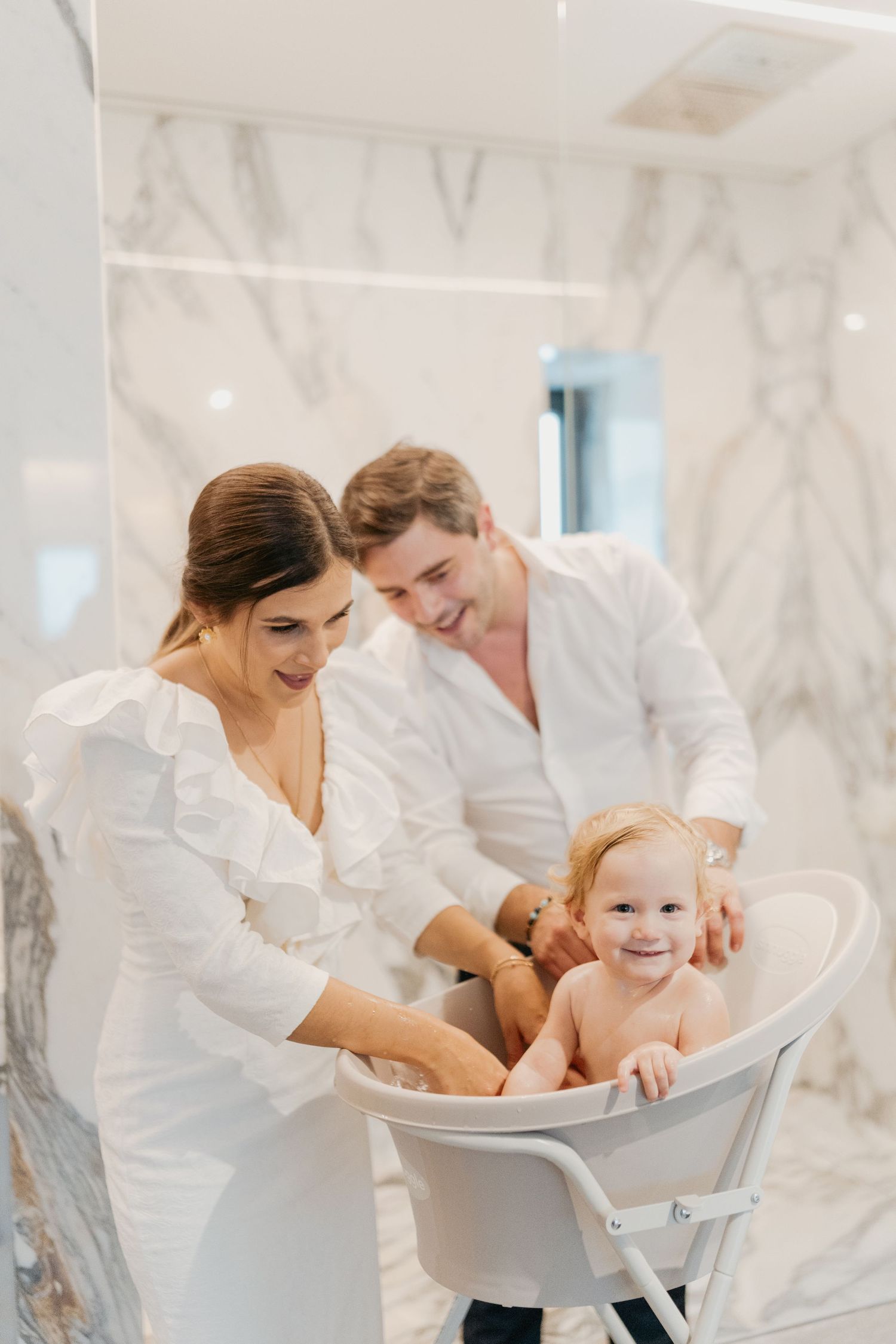 A couple bathes a happy baby in a white marble bathroom during a tender family moment.