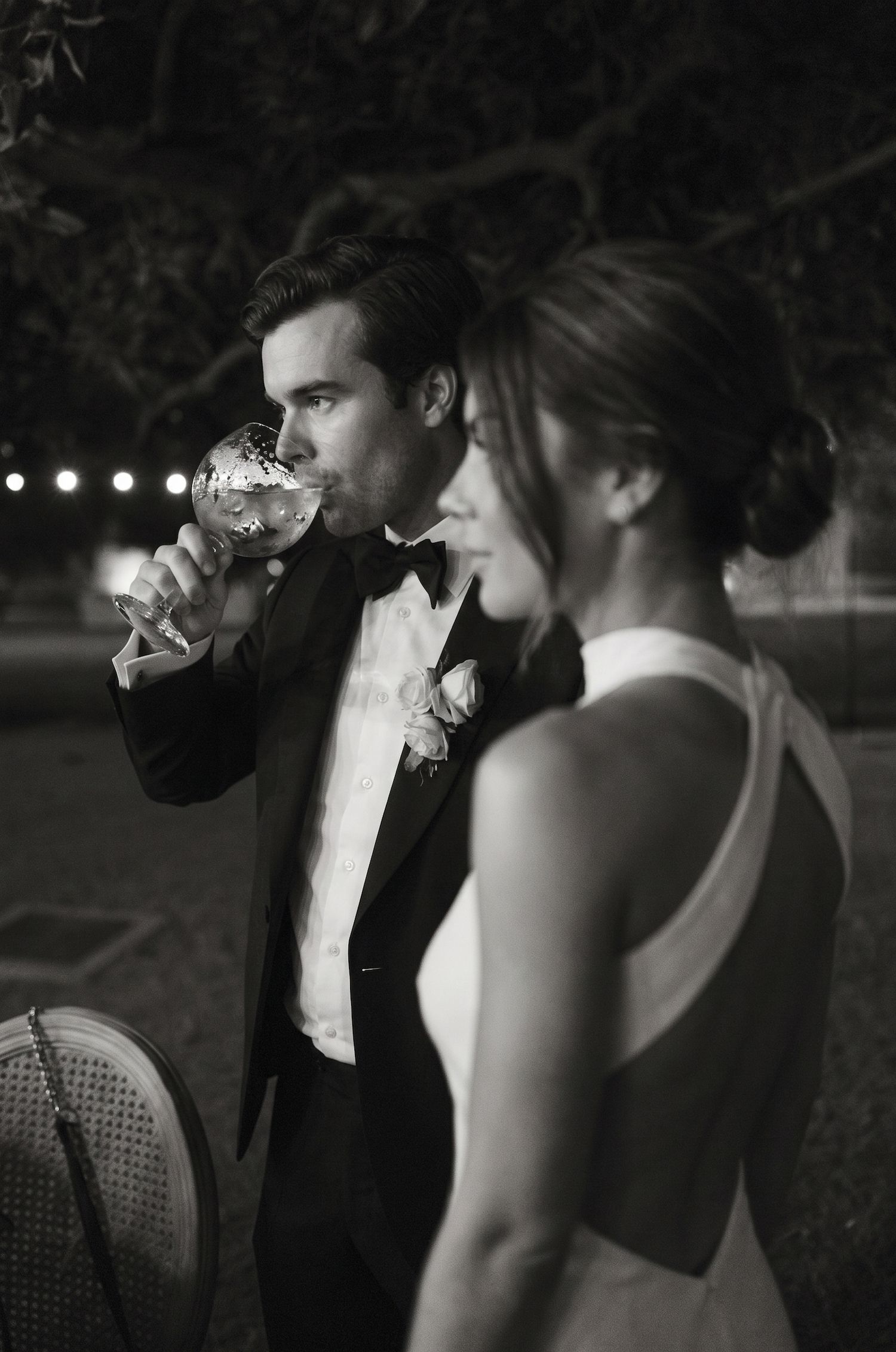 Black and white wedding photo of groom drinking from wine glass beside bride in elegant dress at evening reception.