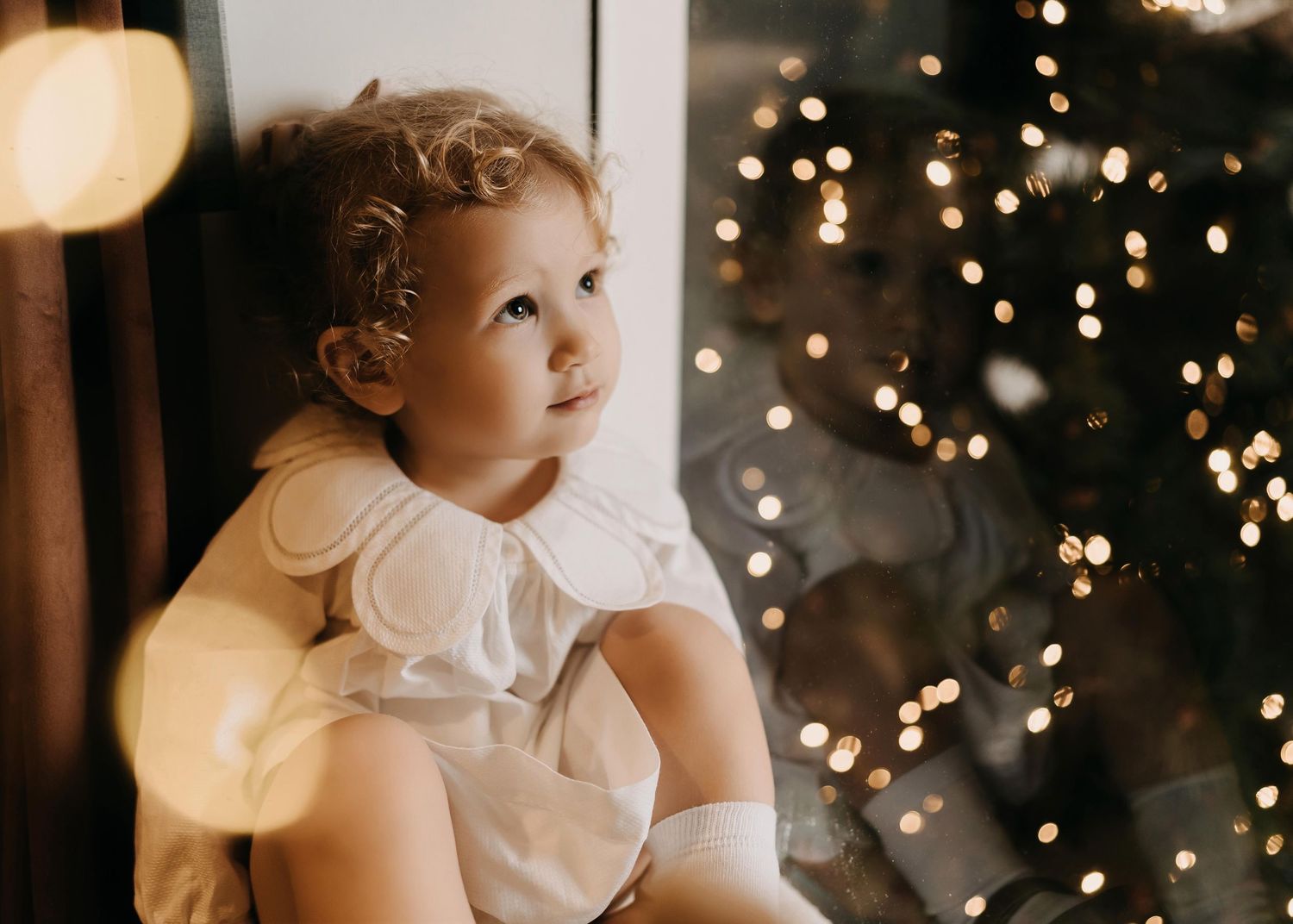 A child in a white dress sits near twinkling Christmas lights creating a dreamy holiday atmosphere.