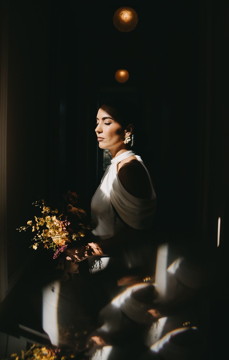 Dramatic side-lit portrait in dark room with autumn-colored floral bouquet creating moody artistic atmosphere.