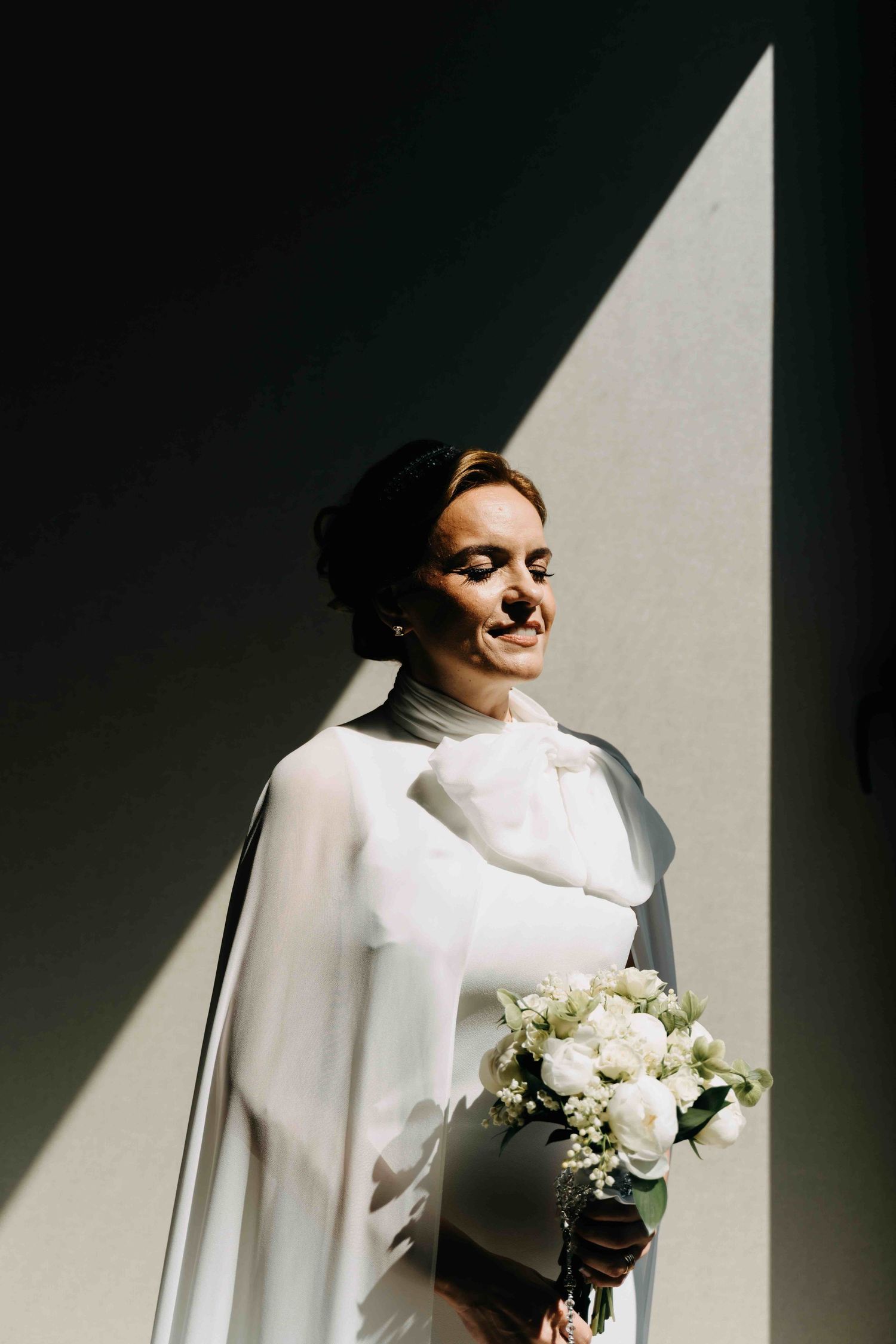 A figure in a white turtleneck dress holds a white floral bouquet while standing in dramatic lighting and shadows.