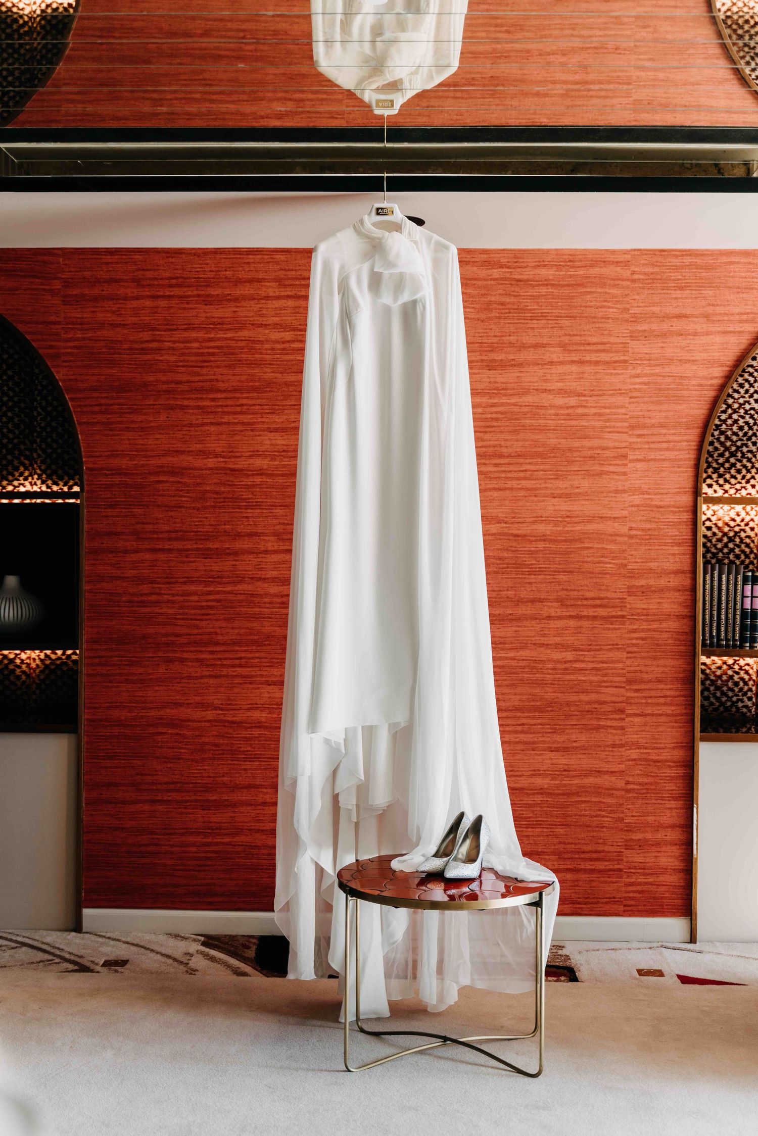 Elegant white wedding gown hangs against coral wall with black heels placed on modern round side table.