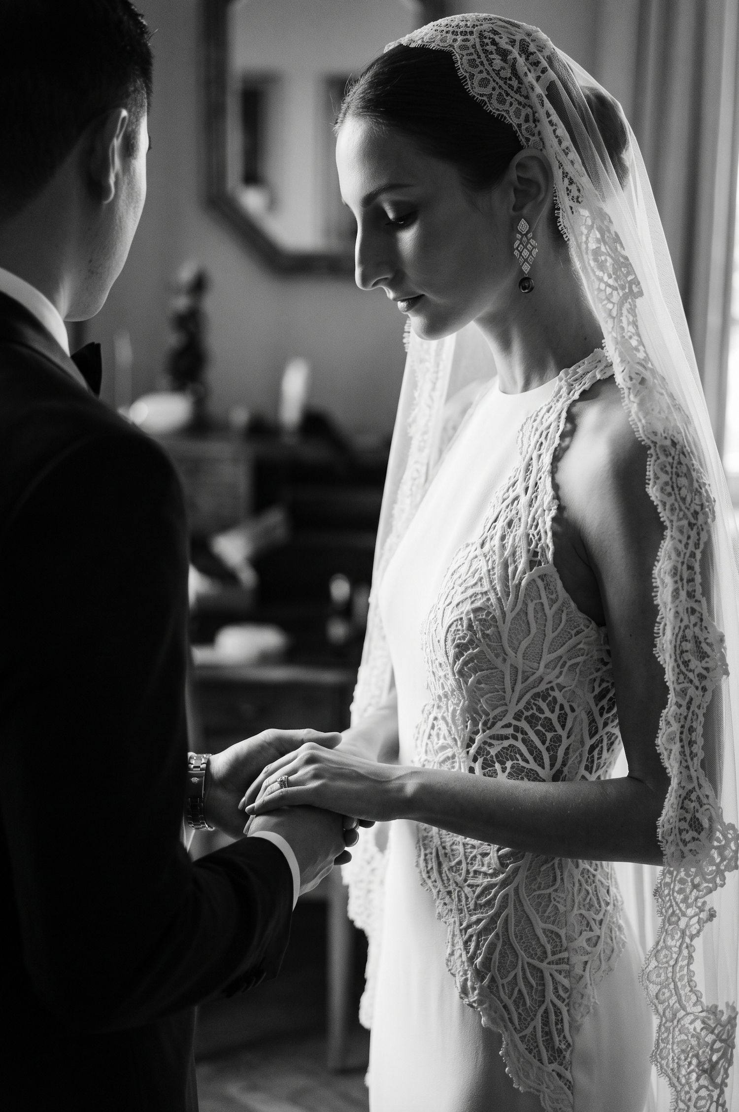 Black and white photo of bride in lace gown and veil exchanging rings with groom during wedding ceremony.