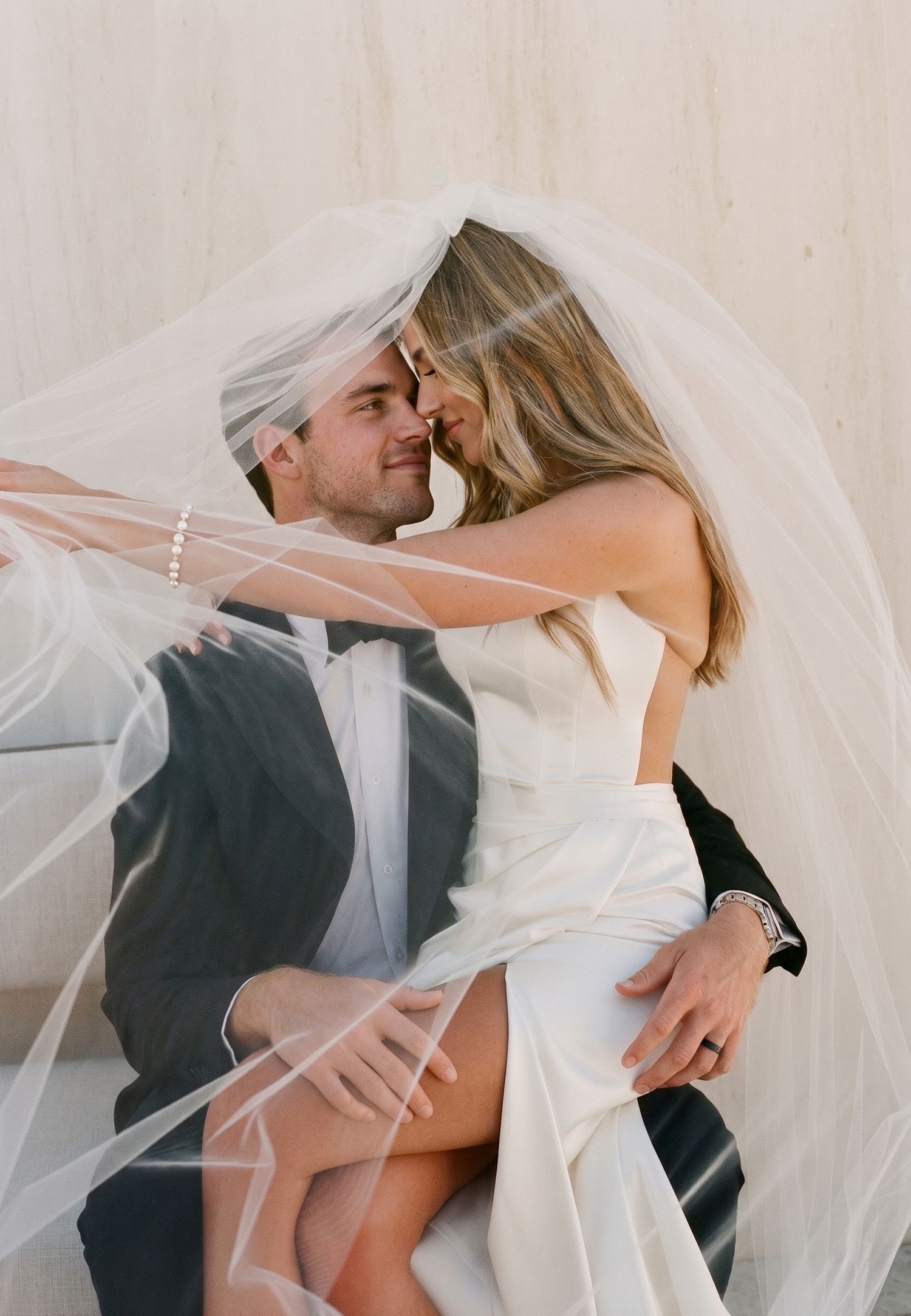 Bride and groom share a romantic moment under a flowing white veil, touching foreheads in an intimate wedding portrait.