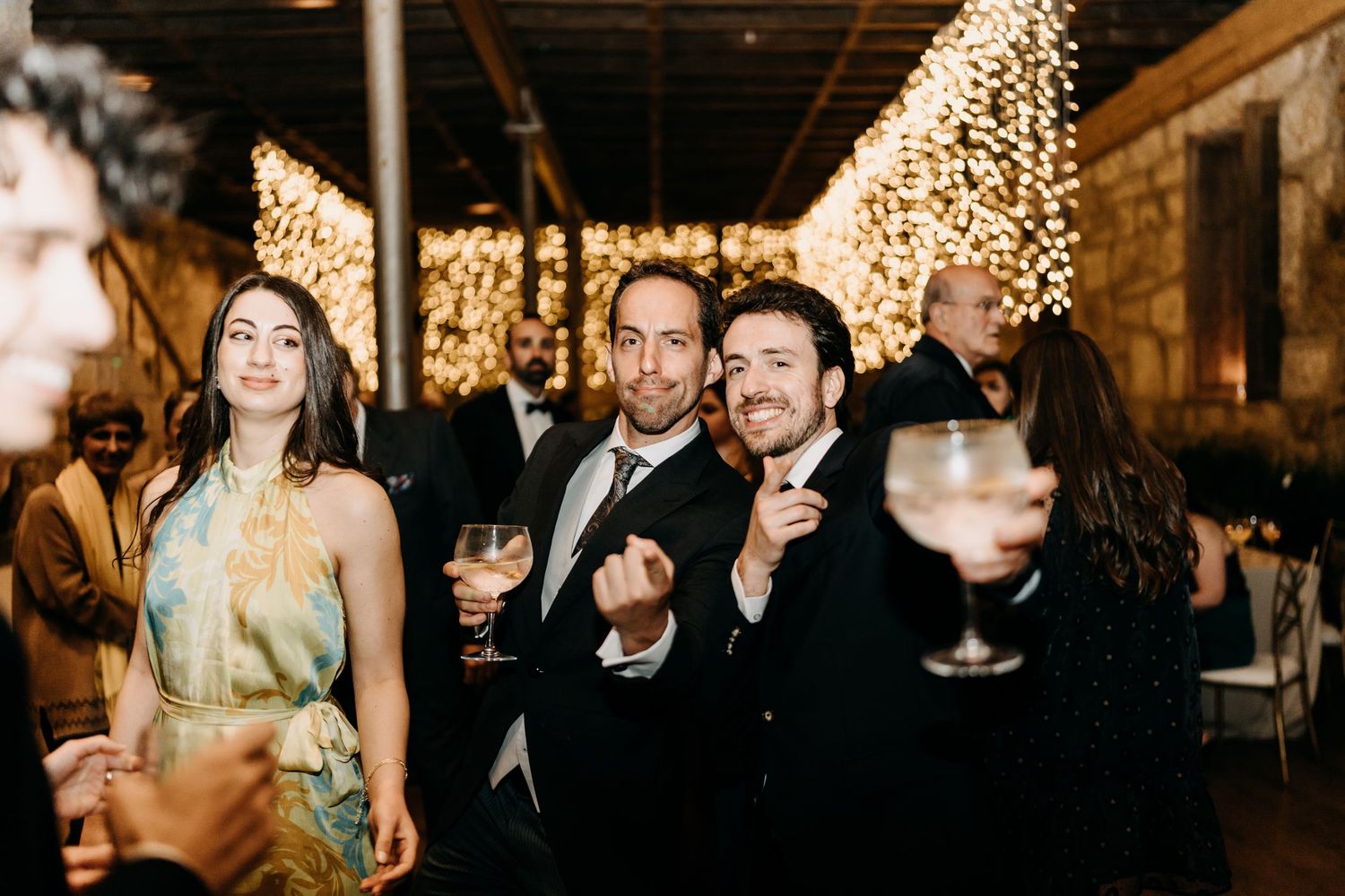 A group of wedding guests dance and celebrate on a dimly lit dance floor illuminated by string lights.