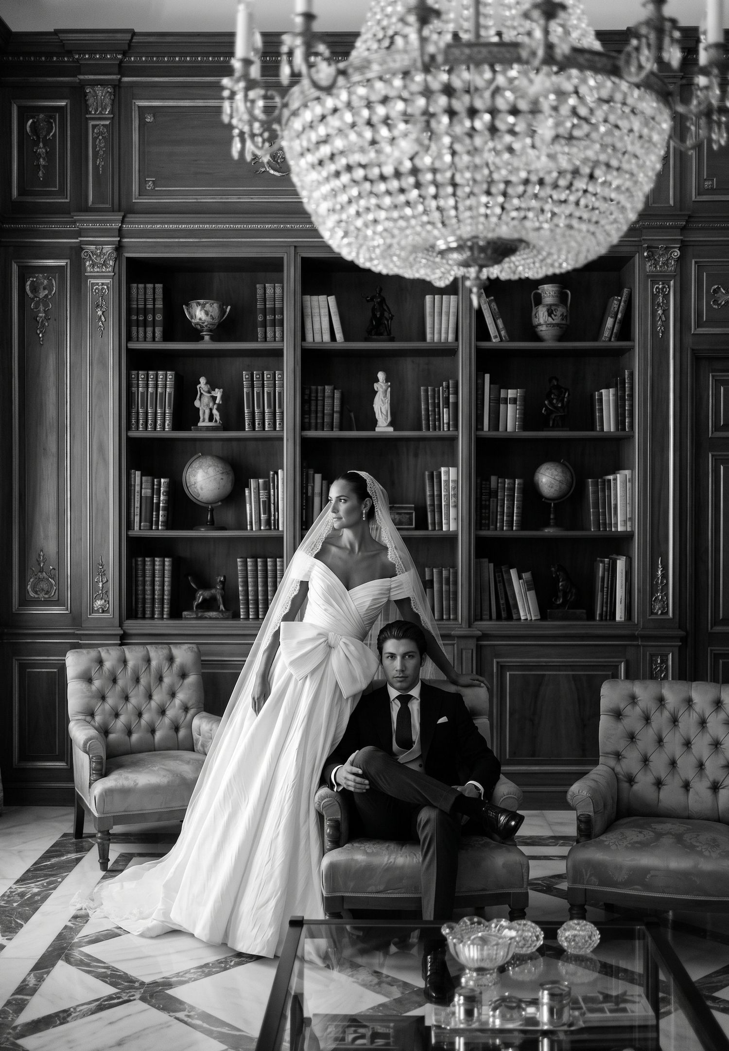 Elegant black and white wedding photo of bride in gown and groom in suit posed in ornate library with crystal chandelier.