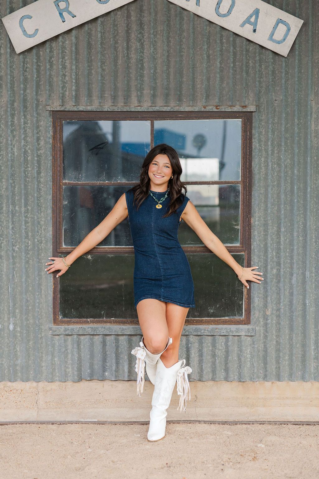 Young woman in blue mini dress and white cowboy boots outside a rustic crossroads building in a photo taken by a Houston senior photographer