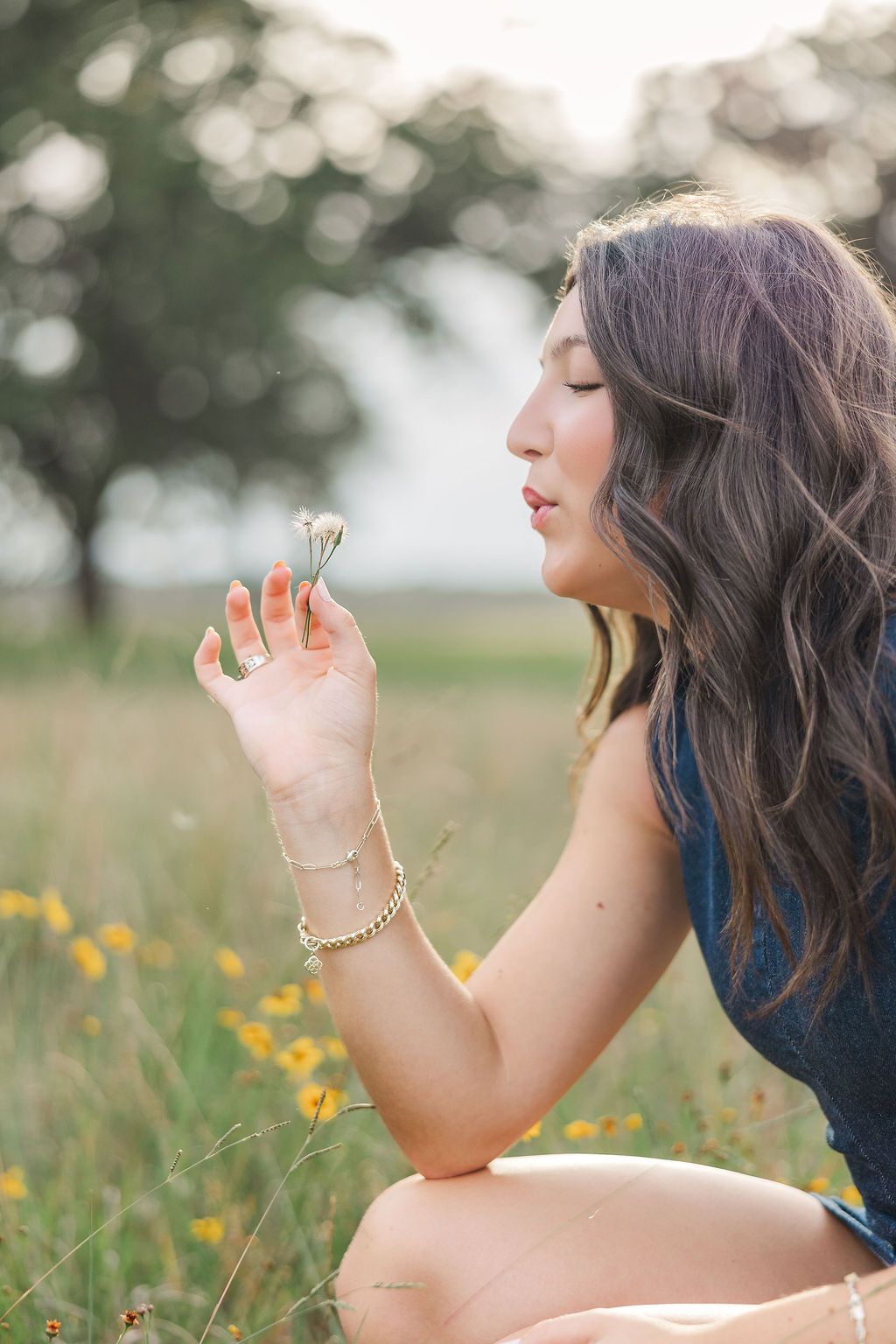 Young woman with wavy hair blowing a dandelion in a sunny wildflower meadow, wearing a blue dress and gold bracelet.