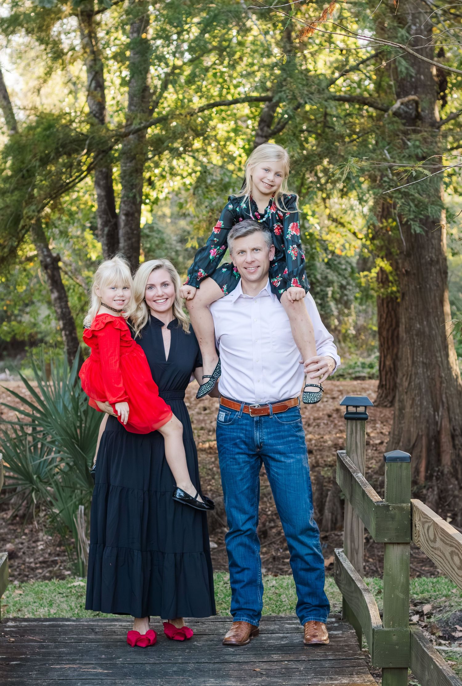 A family of four posing on a wooden bridge surrounded by lush green trees, wearing festive holiday outfits for houston christmas pictures.