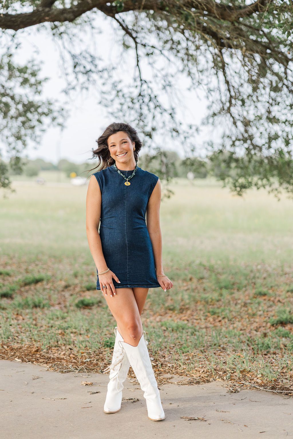Houston high school senior girl in denim mini dress and white cowboy boots smiling outdoors under a tree.