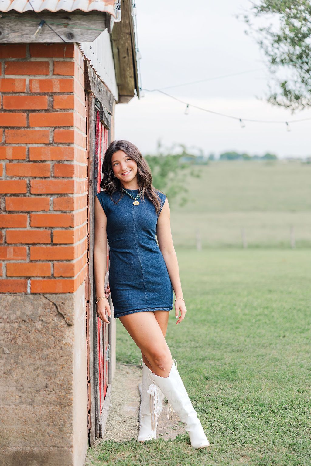 Young woman in denim mini dress and white cowboy boots leaning against rustic brick building on green farm.