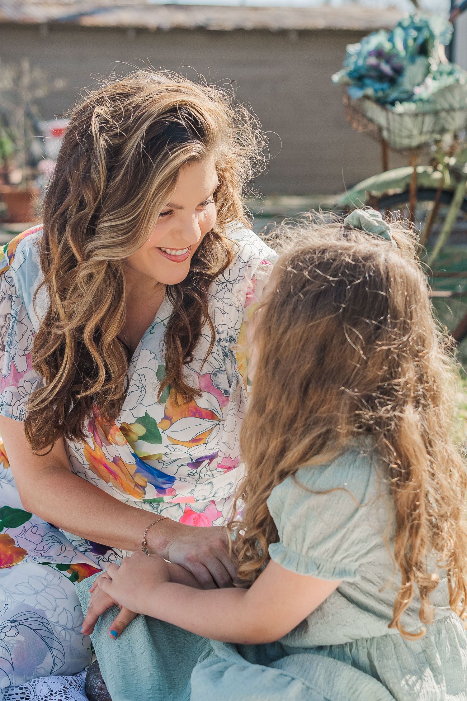 A smiling woman with wavy hair in a floral dress sits outdoors with her young daughter during a session with Houston family photographer.
