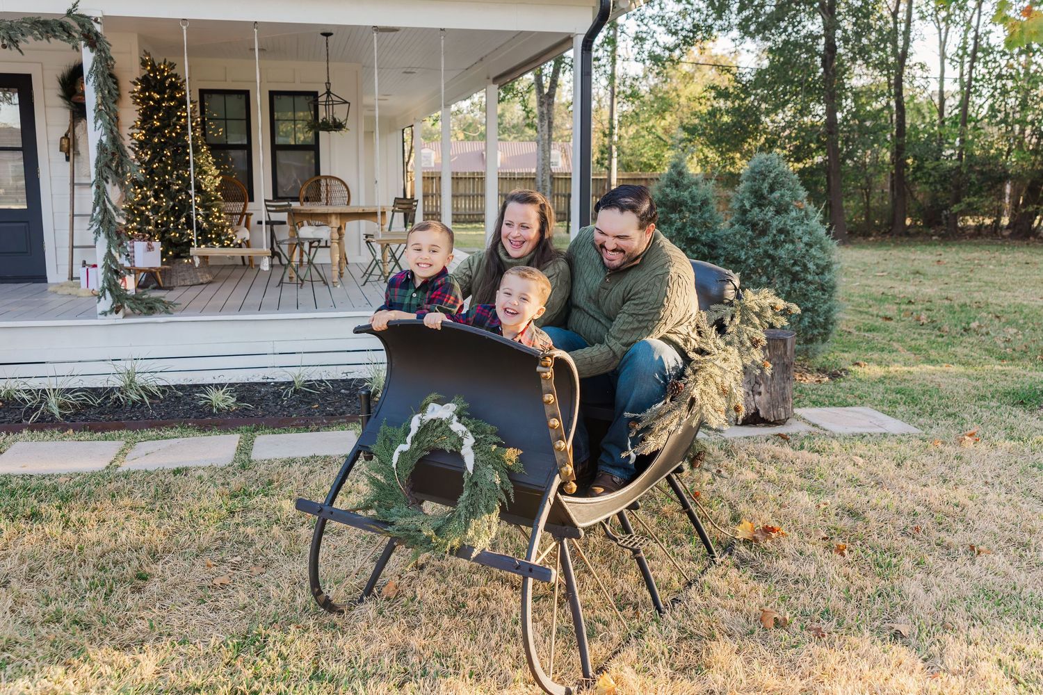 Family of four joyfully sitting in a vintage sleigh outdoors near a holiday-decorated porch during Houston christmas pictures.