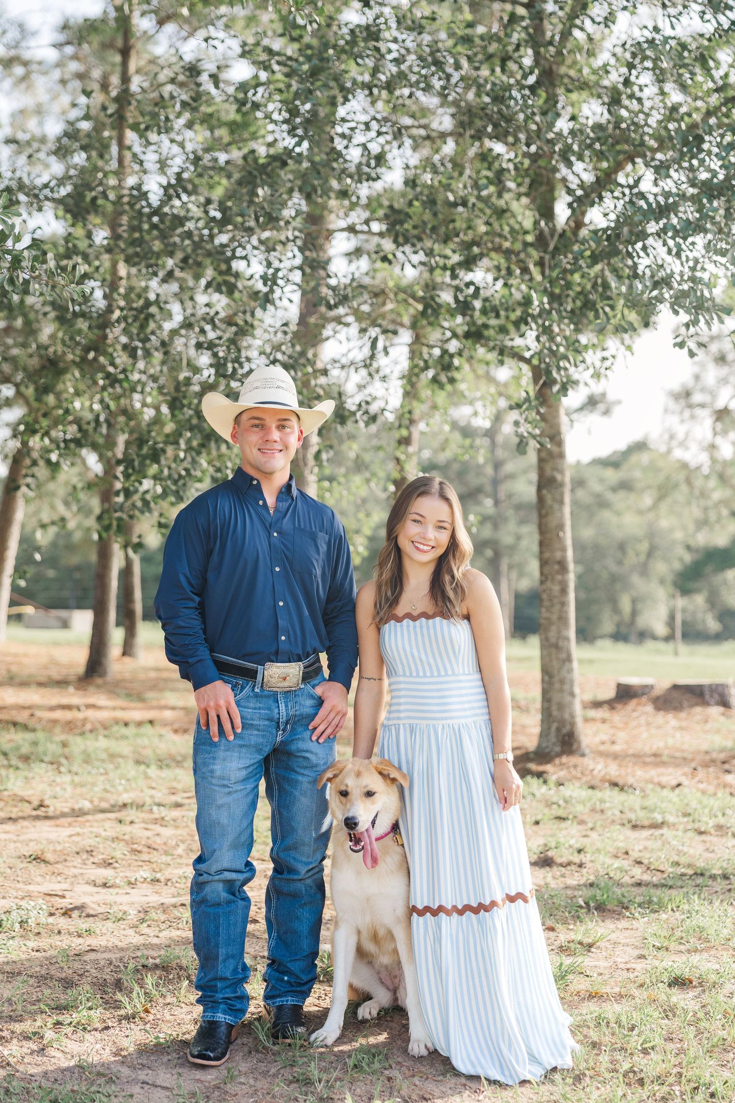 A smiling couple in casual attire poses with their golden dog outdoors among tall trees on a sunny day during family photos in Houston TX.
