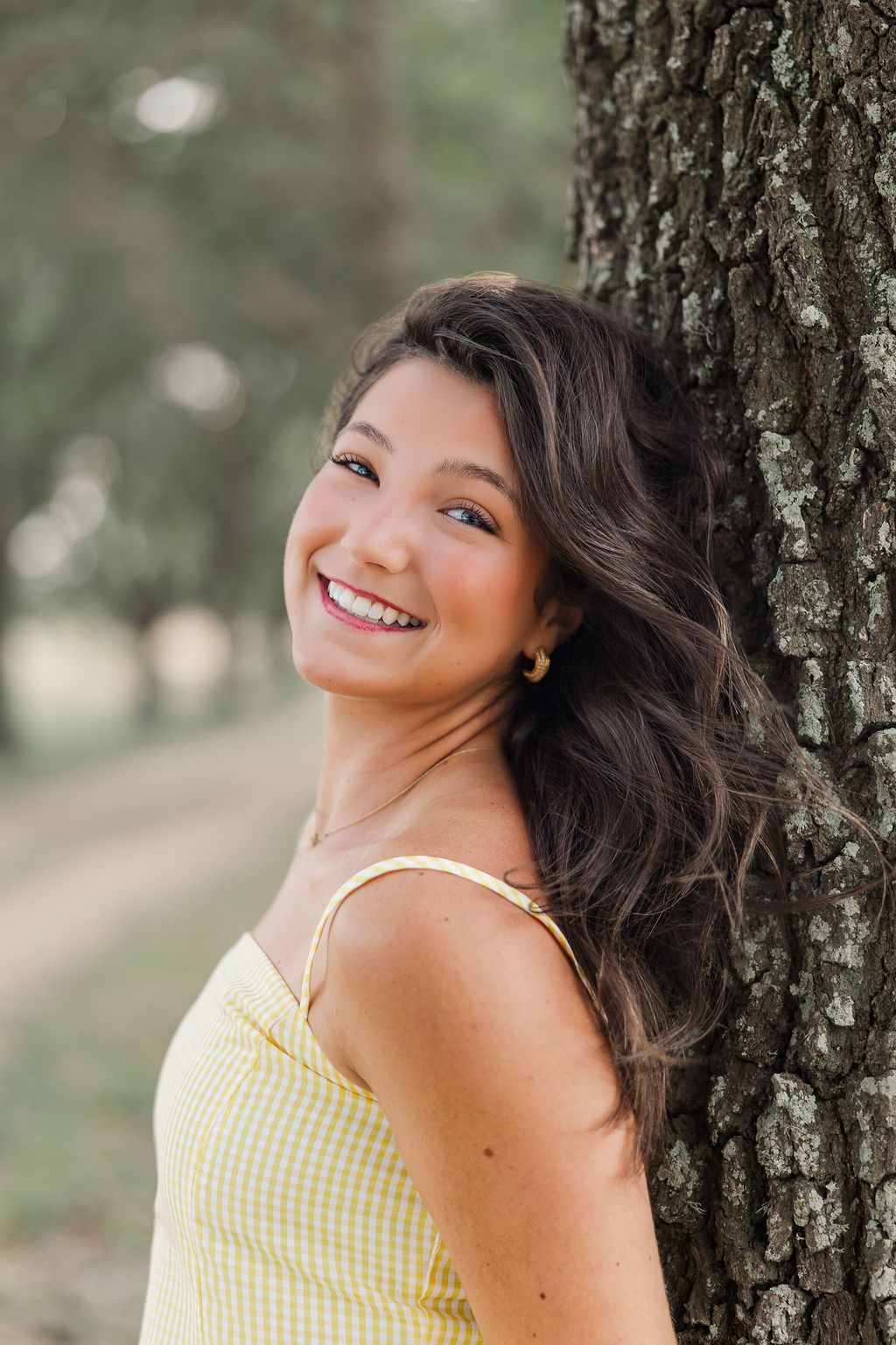 Young woman with dark wavy hair smiling outdoors, wearing a yellow gingham dress while leaning against a tree for Houston senior photos.