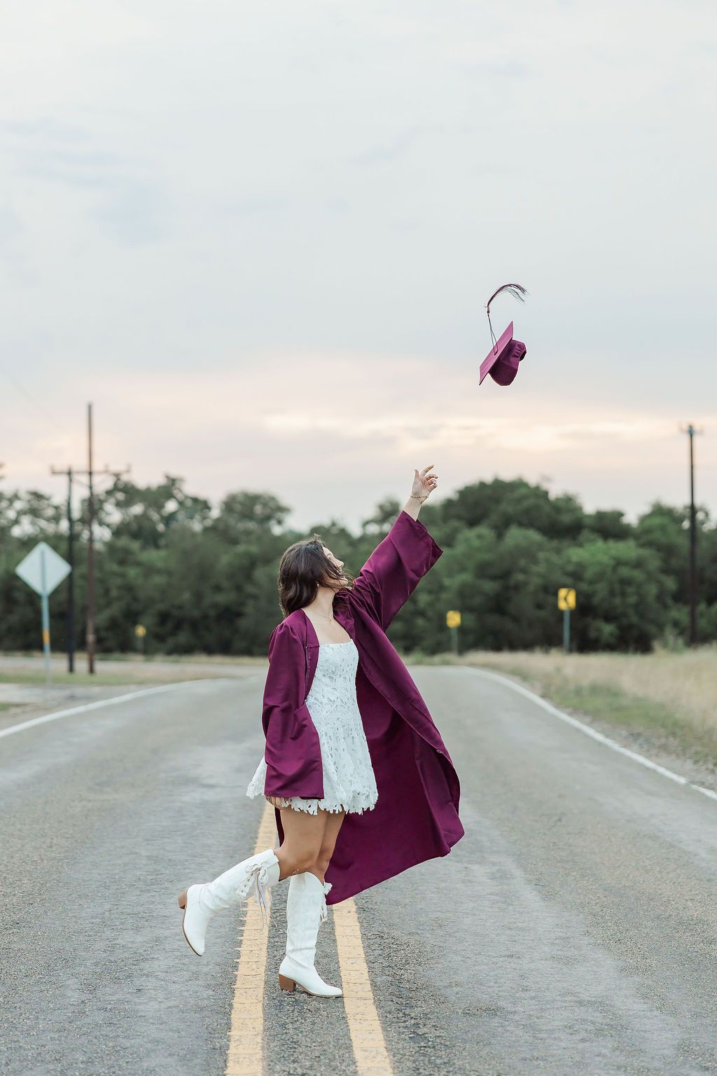 Graduate in maroon gown tosses cap on open road, celebrating achievement in a scenic outdoor setting.