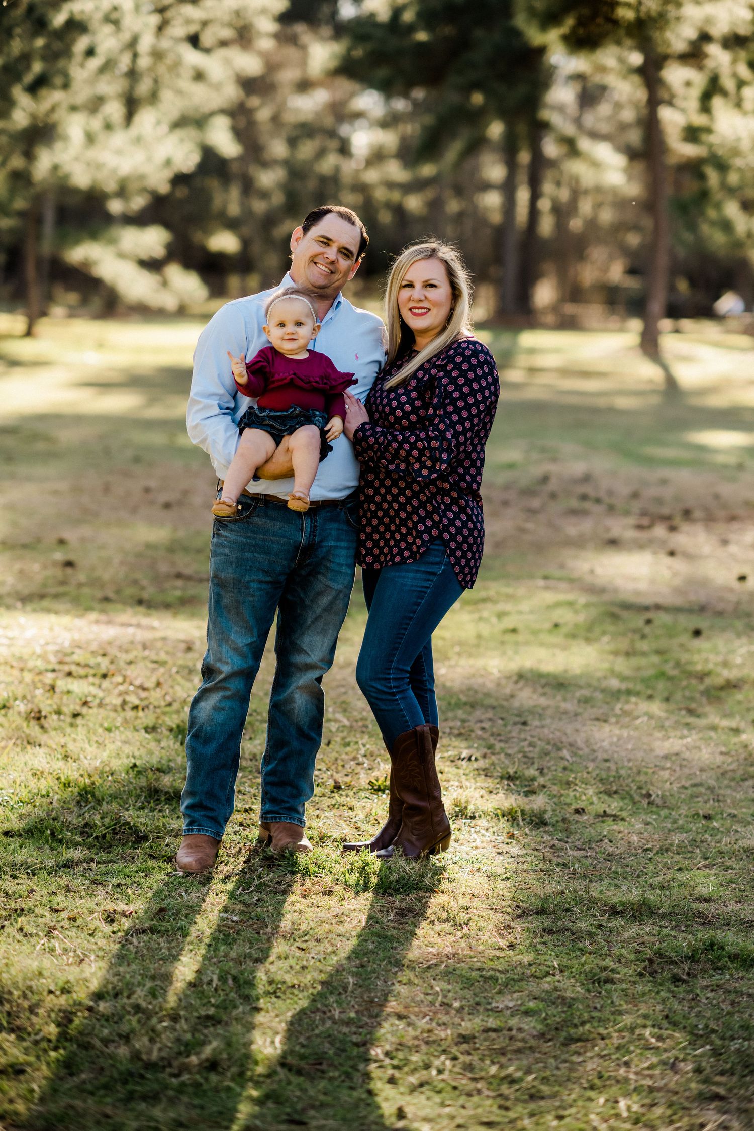 A family of three poses together in a sunlit wooded area wearing casual clothing.