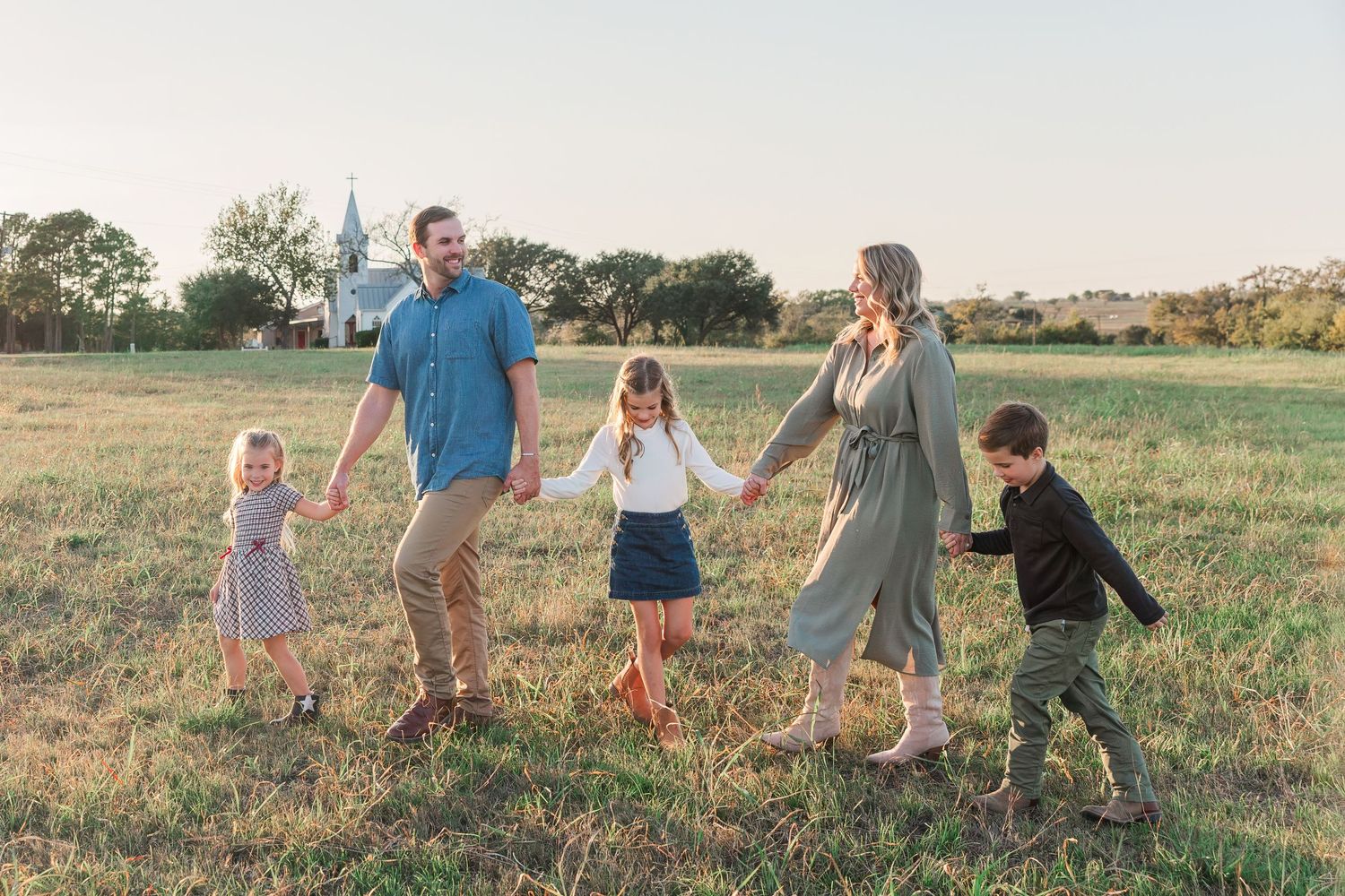 Family of five holding hands walking through a sunlit field with a white church in the background.