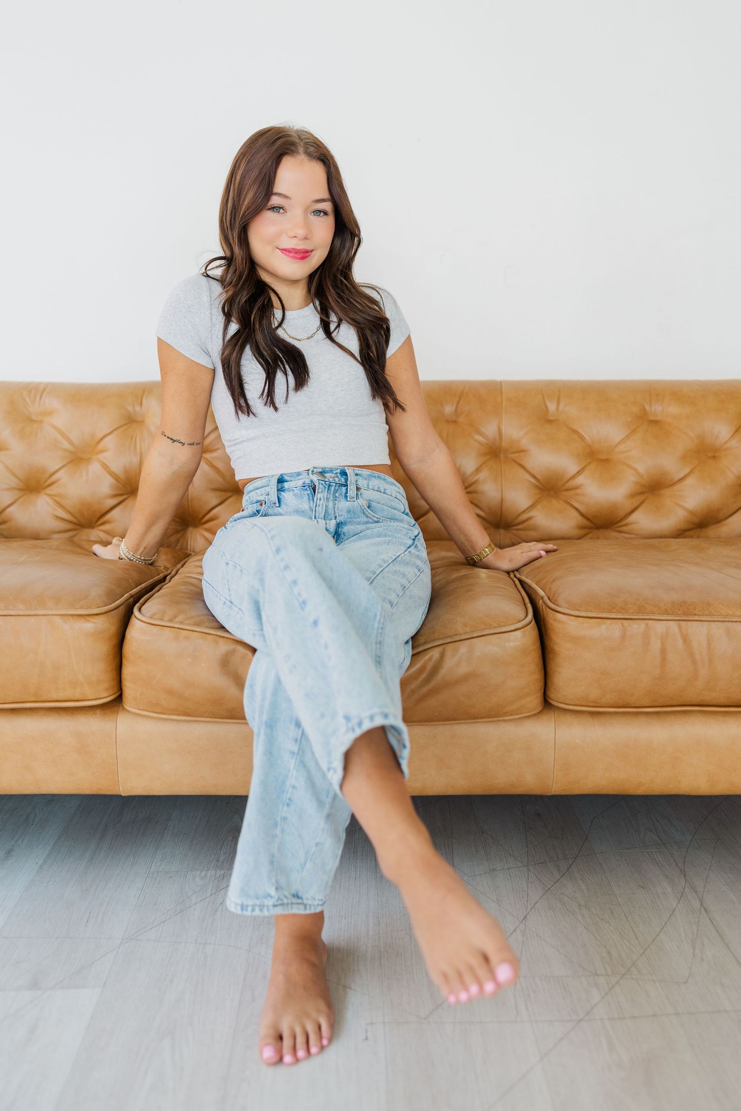 Young woman with dark hair sits barefoot on a tan tufted leather sofa during Houston senior studio portaits.