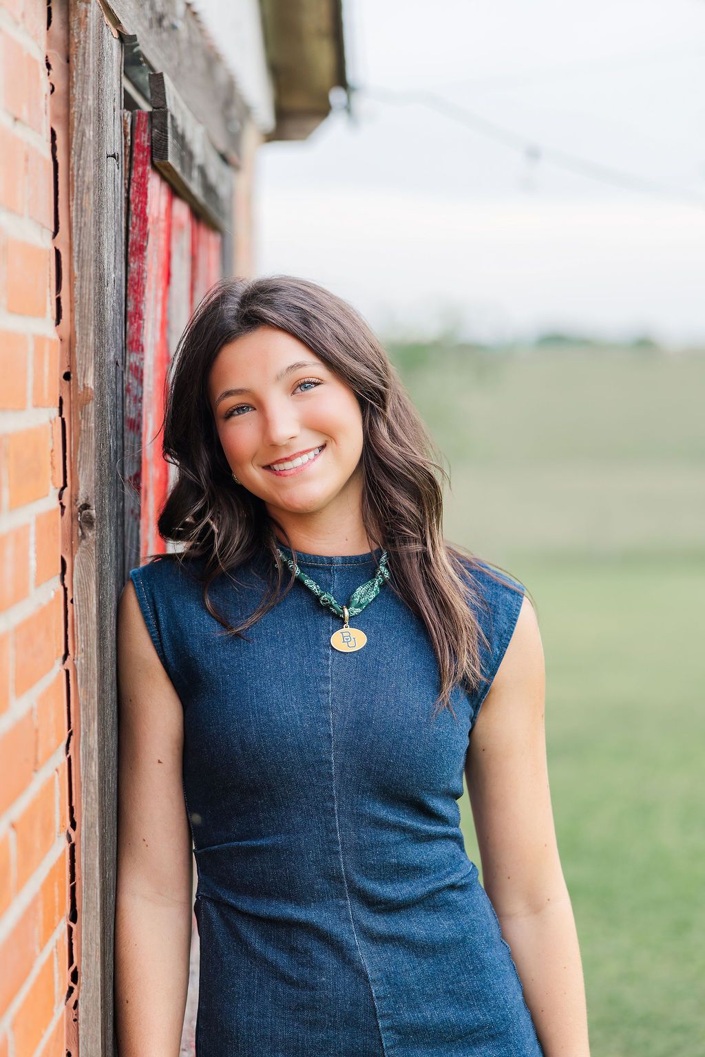 Young woman in denim dress smiles leaning against rustic brick wall outdoors during a Houston senior photoshoot.