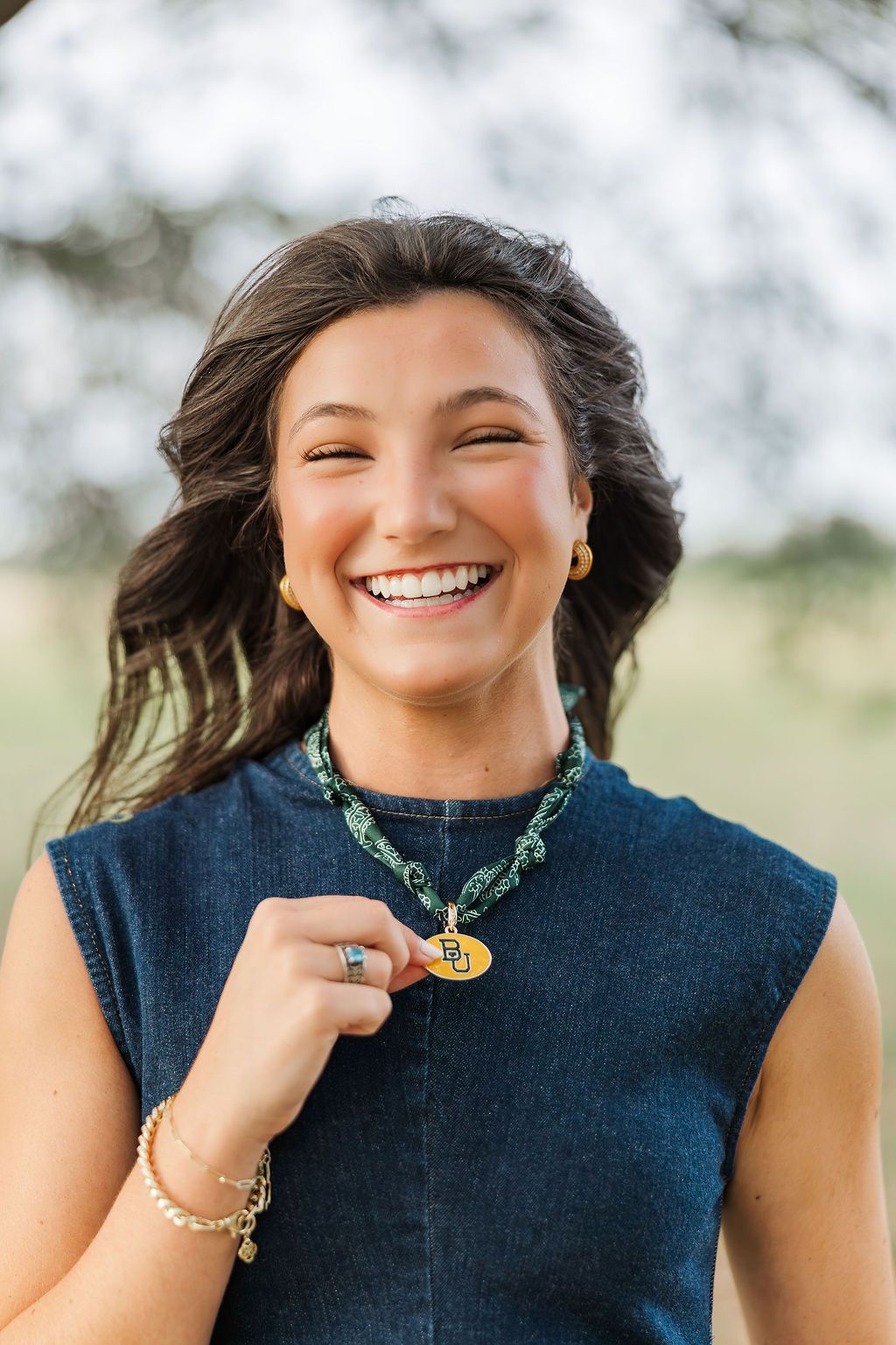 Young woman laughing joyfully outdoors, wearing denim top and green beaded necklace with gold pendant.