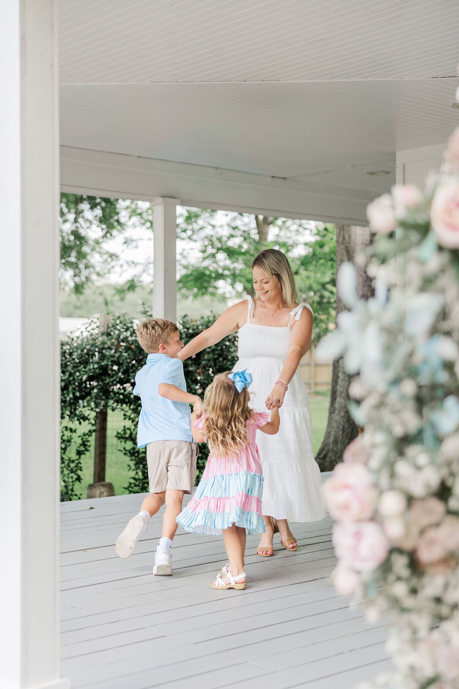 A mother in a white dress dances with her two young children during a motherhood session with a Houston newborn photographer.