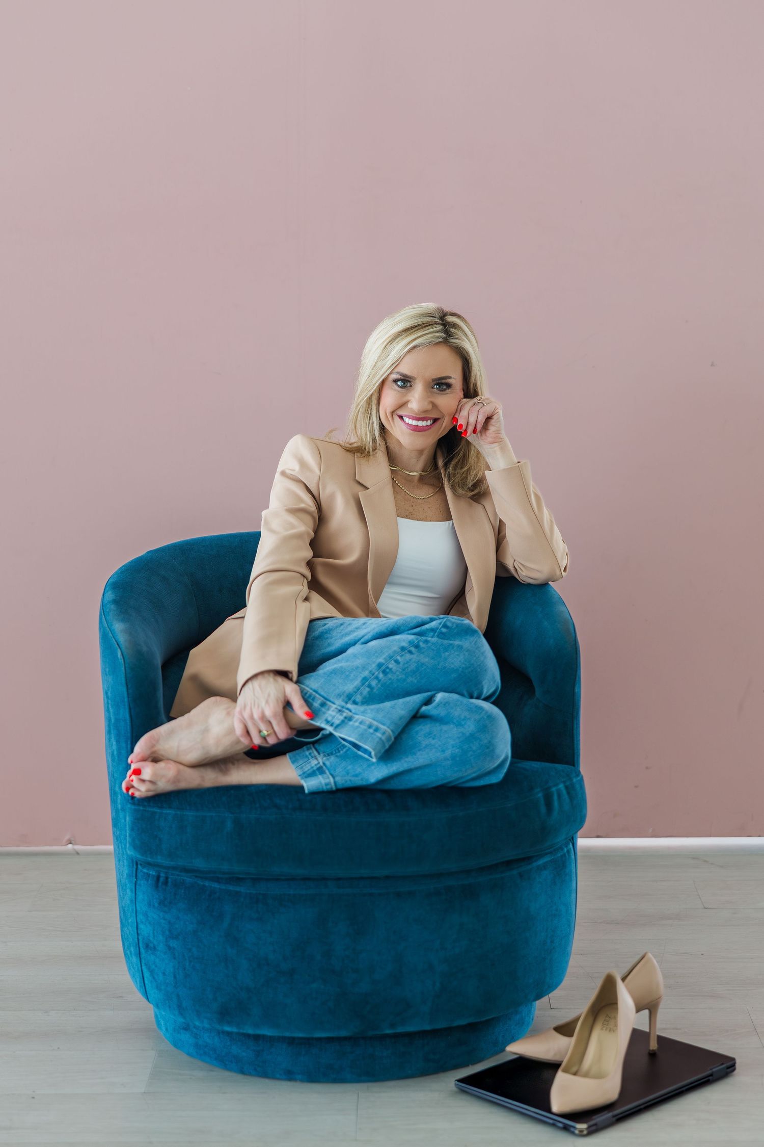 Smiling blonde woman in beige blazer and jeans sitting cross-legged in blue velvet chair with heels on floor.