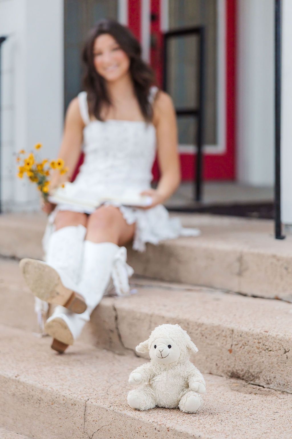 A small lamb stuffed animal sits on stone steps in front of a smiling young woman in a white dress holding flowers.