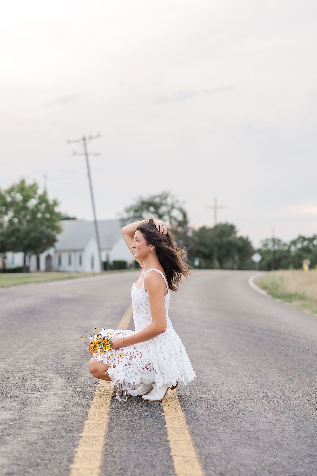 Woman in white lace dress crouching on a rural road holding wildflowers, hair blowing in the wind.