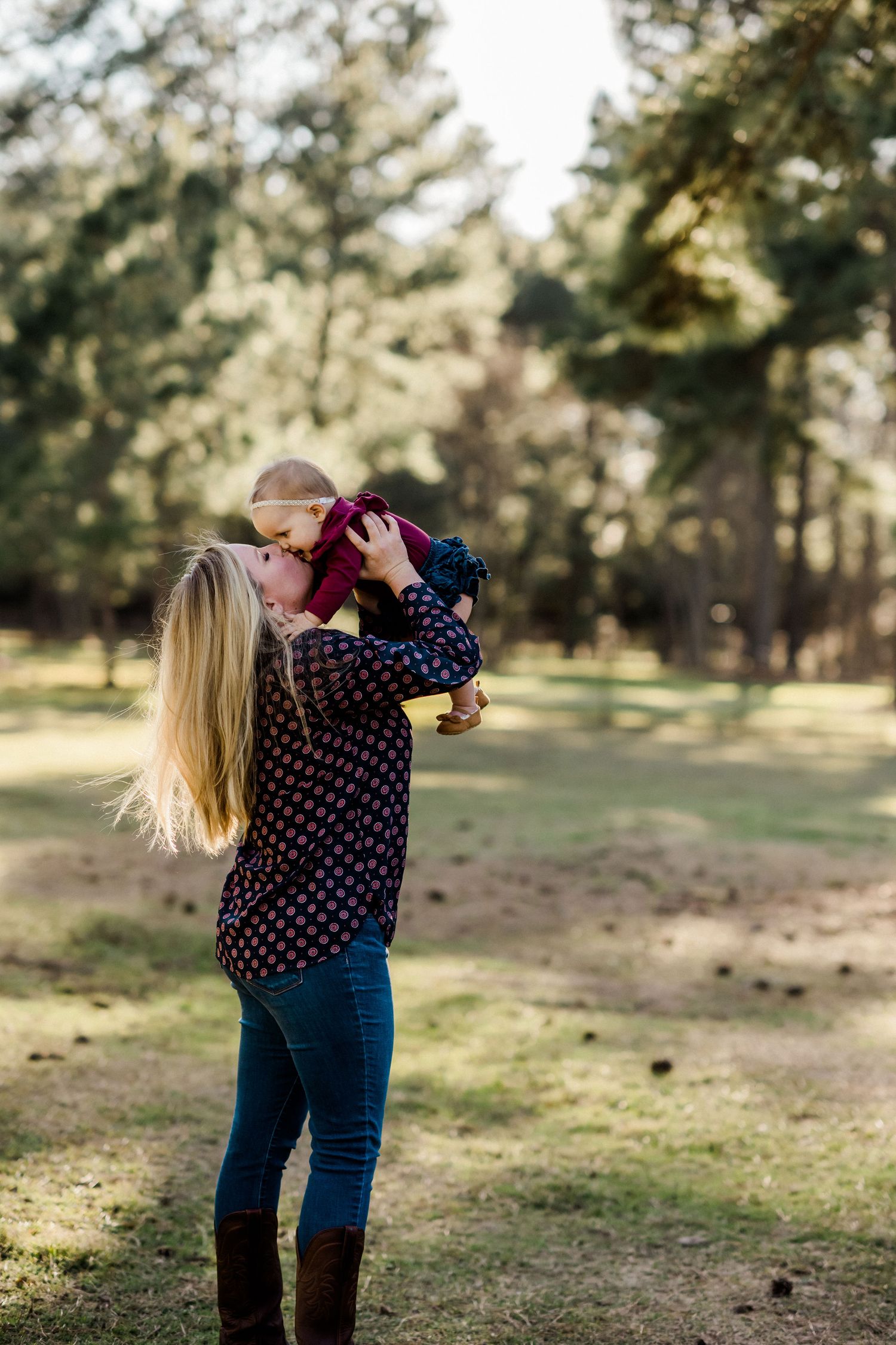 A woman in a polka dot shirt holds up a small child in a wooded outdoor setting.