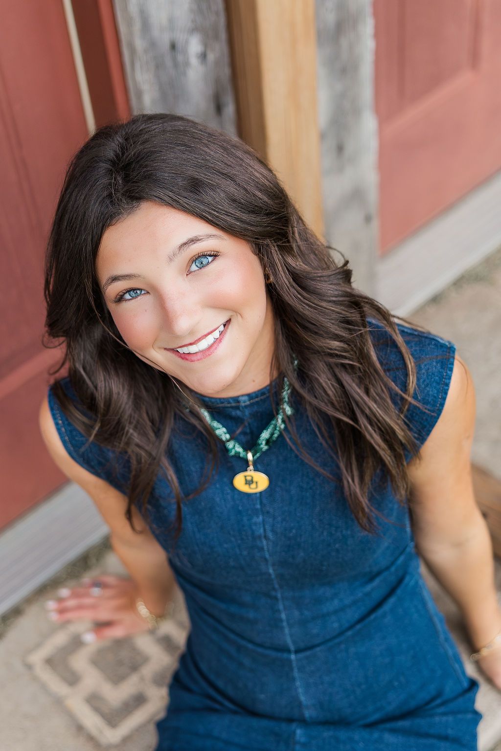 Smiling brunette woman with blue eyes wearing a denim dress and turquoise necklace seated outdoors.