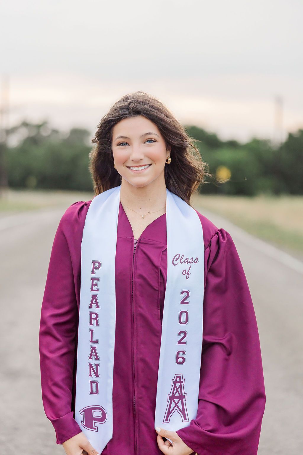 Smiling young woman in maroon graduation gown and Pearland Class of 2026 sash poses on a road outdoors.