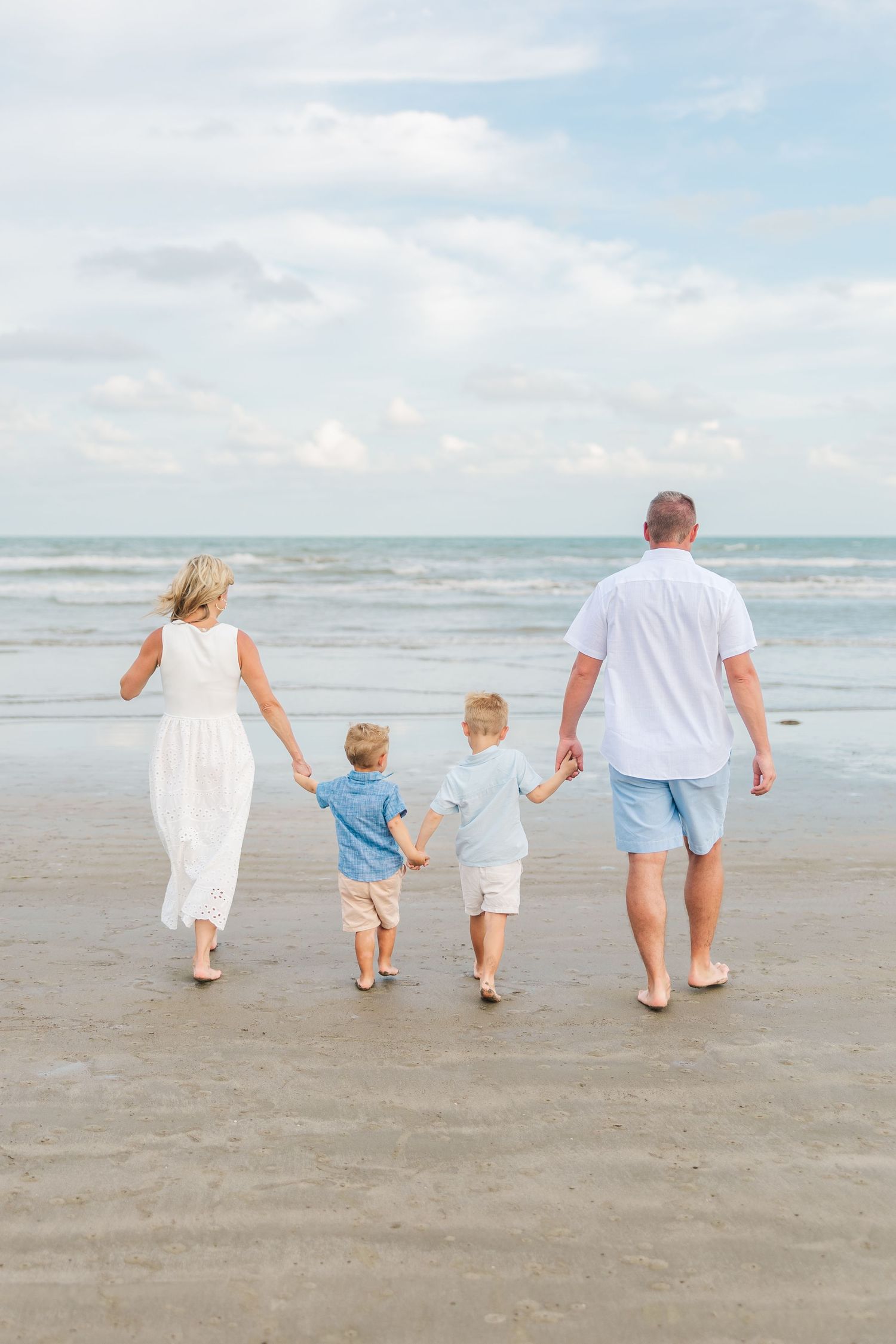 Family of four walking hand-in-hand on a sandy beach toward ocean waves on a cloudy day in a photo taken by a Houston family photographer.