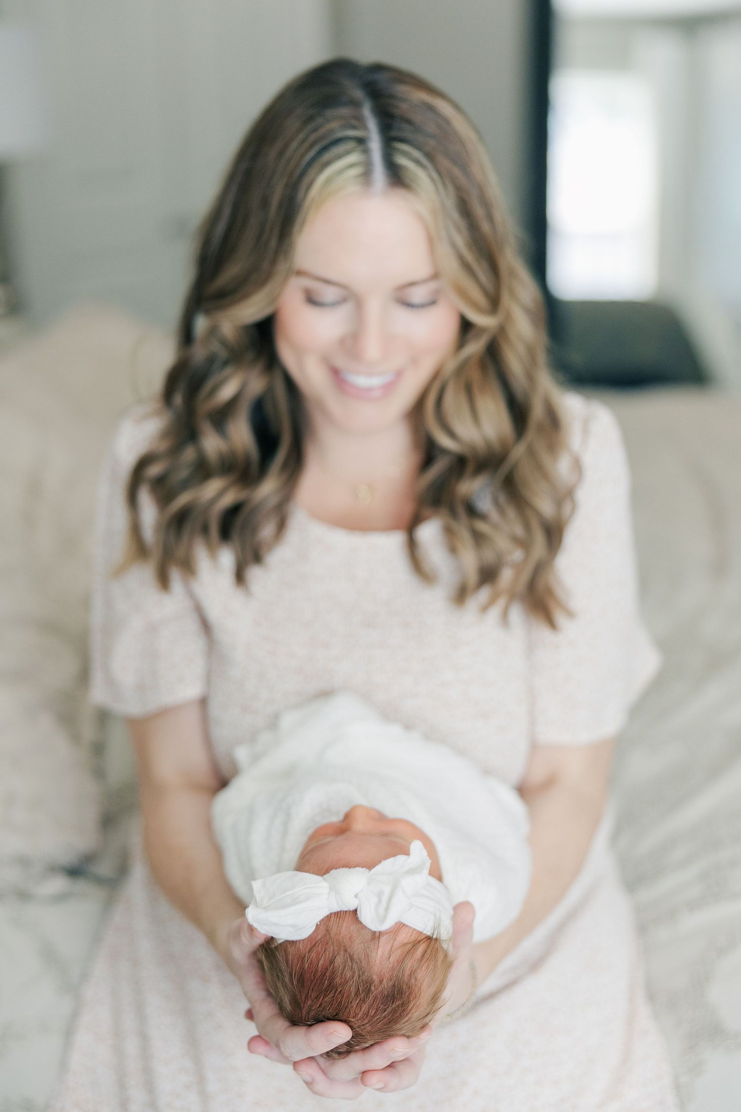 Smiling mother in beige dress tenderly holds swaddled newborn baby in a photo taken during a Houston newborn photography session.