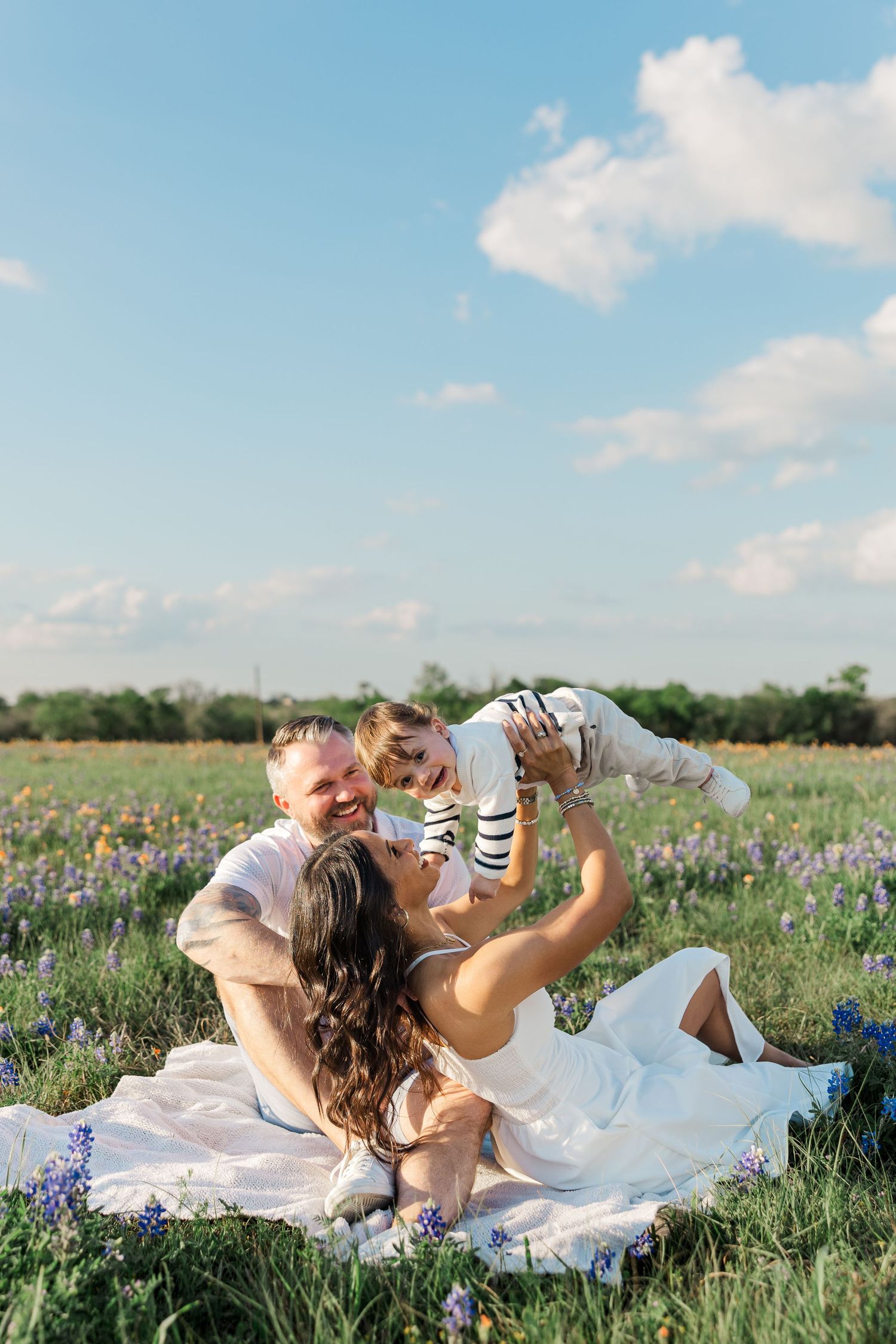 A family sitting in a field, parents lifting smiling toddler under blue sky during a photoshoot with a Houston family photographer.