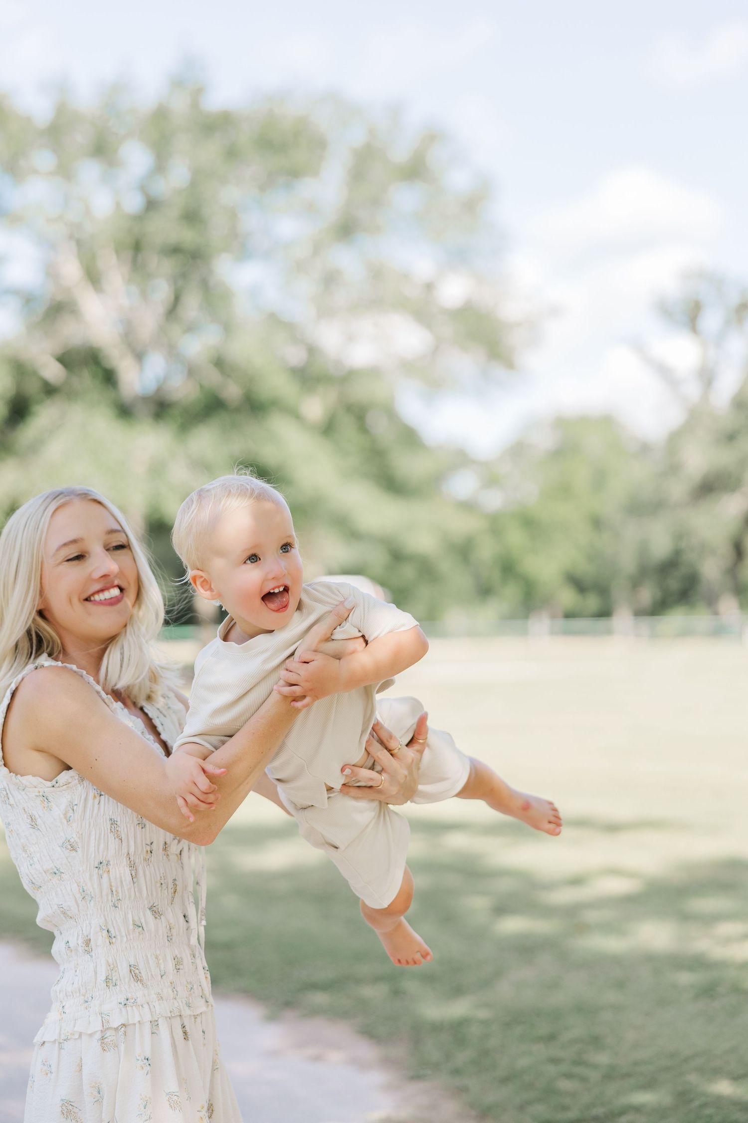 A mother lifts a happy baby outdoors in a lush green park on a sunny day during photography in Houston.