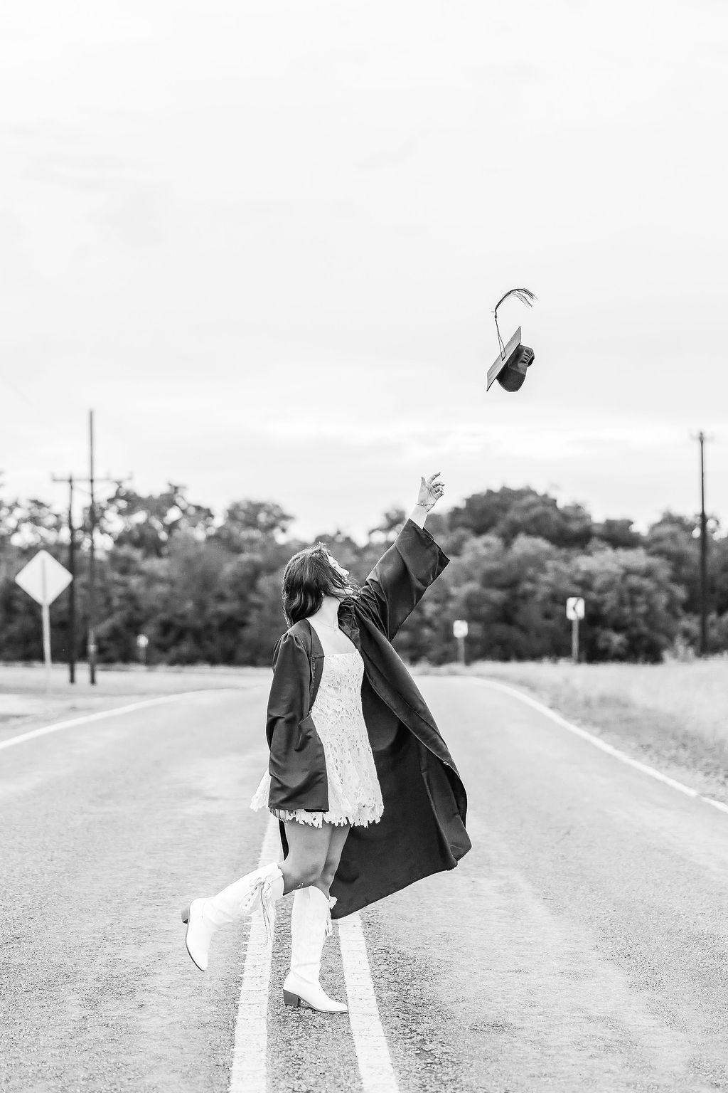 Black and white photo of a graduate tossing cap in air on empty road, wearing gown and boots.