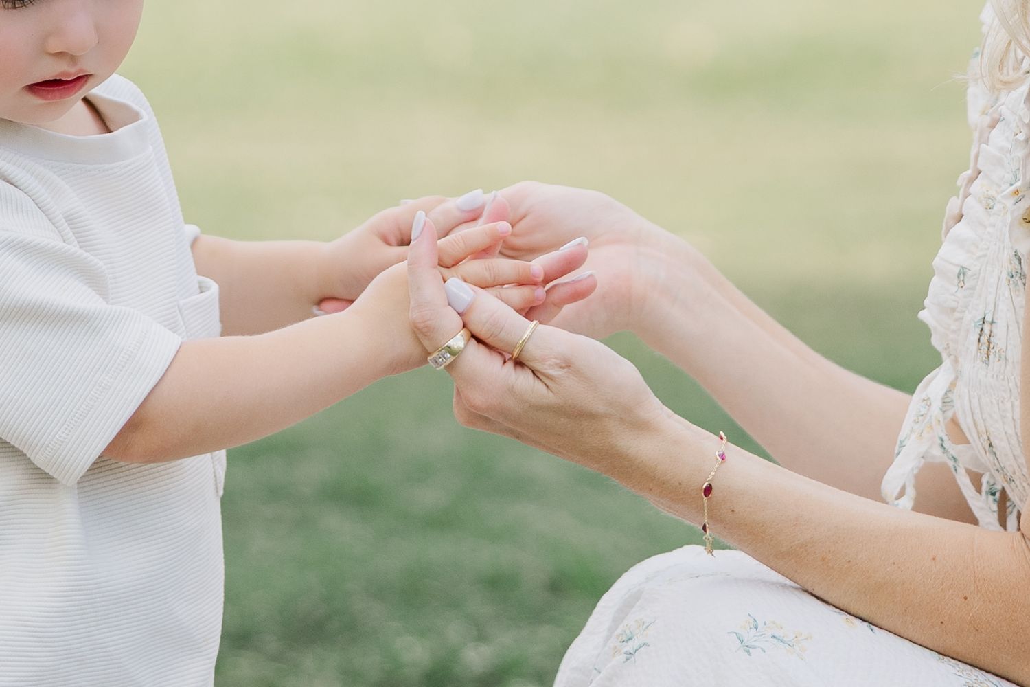 A mother gently holds her toddler's hands outdoors on a green lawn, wearing a bracelet and floral dress.