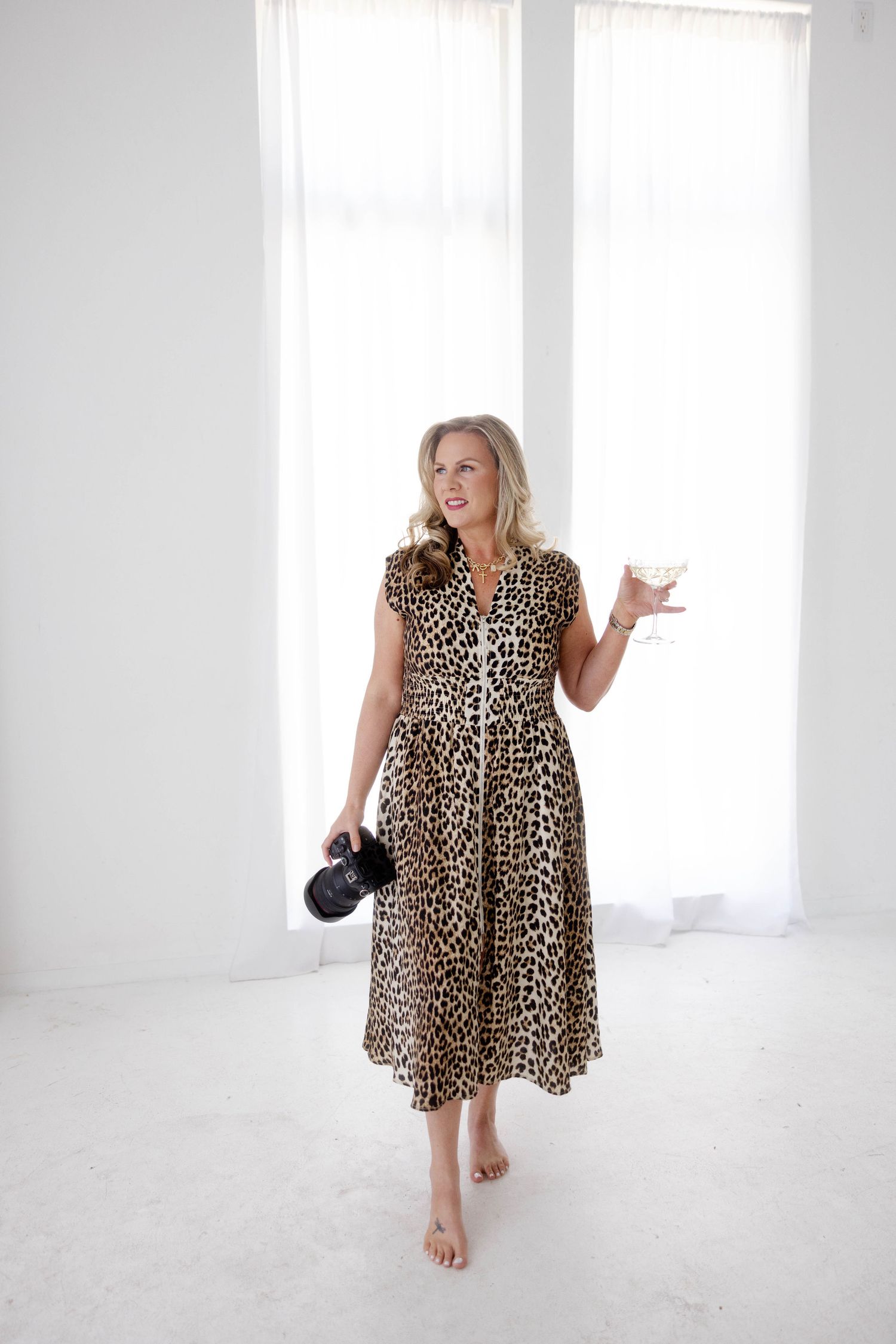 A Houston senior photographer in leopard print midi dress holding camera and martini glass, standing barefoot in bright white room.